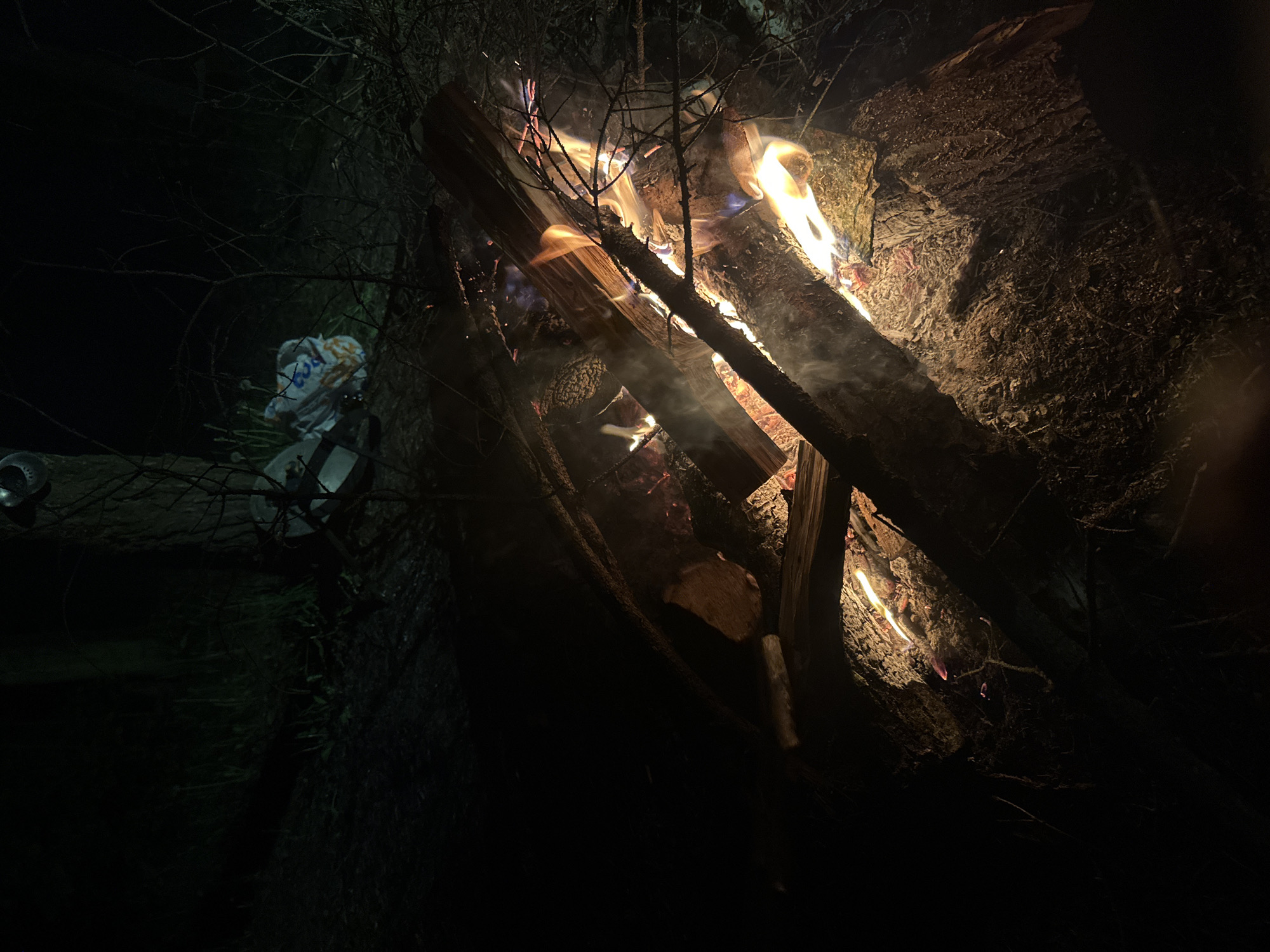 The image shows a nighttime scene in a wooded area of Юзуфоўскі сельскі Савет, Belarus. At the center is a campfire burning with logs and smaller branches. The flames are visible, illuminating the wood and creating smoke. To the left of the fire, a white plastic bag is placed along with some pieces of metallic cooking/eating utensils, potentially indicating a campsite. The environment is dark, with the surrounding forest trees and undergrowth barely visible.