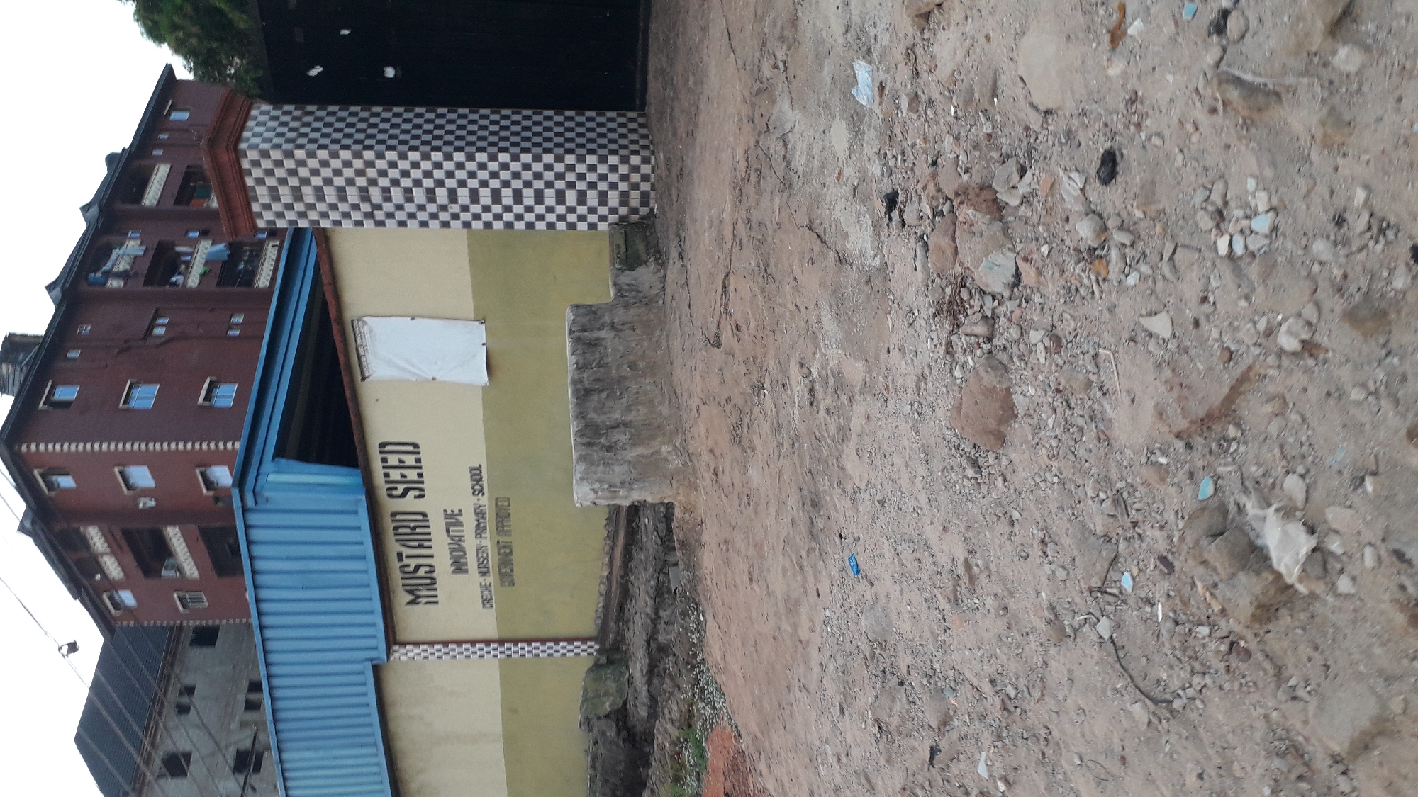 The image depicts the exterior of "Mustard Seed Innovative Creche-Nursery-Primary School, Government Approved," located in Nkpor, Nigeria. The school building has a beige-colored facade with black lettering displaying the school's name. Above the school sign is a blue corrugated metal overhang. To the left, a multi-story residential building with reddish-brown brickwork and blue-tinted windows is visible. Adjacent to the school is a checkered column. In the foreground, a dirt and gravel-covered ground slopes upward.