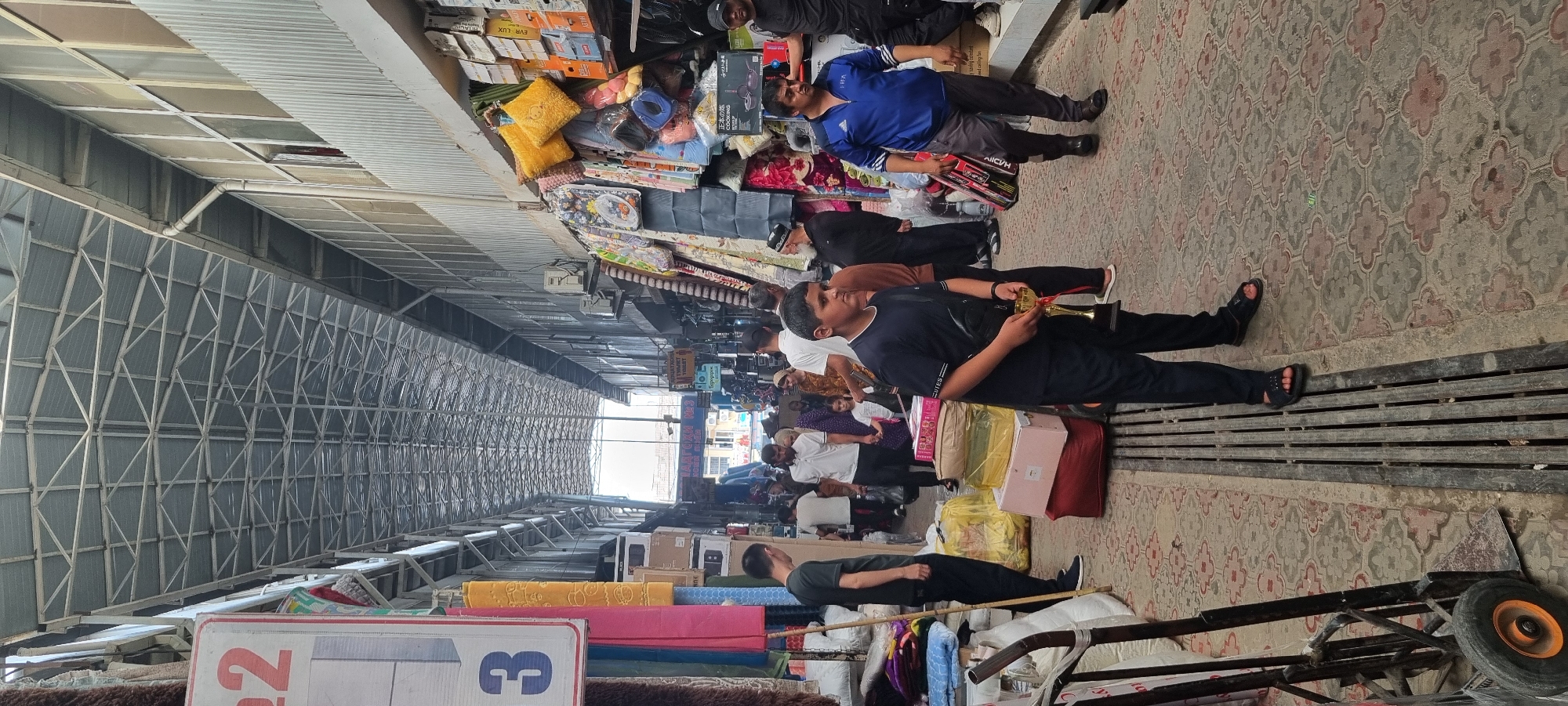 The image depicts an indoor market scene in Bokhtar, Tajikistan. The market appears to be a covered bazaar with a metallic, arched roof structure overhead. The narrow walkway is lined with stalls displaying various goods, including textiles, bedding, and boxed items. People are seen walking through the market, some carrying shopping bags and others interacting with vendors.
In the foreground, a young boy in a dark blue t-shirt and pants is holding a golden trophy. To his left, a trolley with wheels is parked. Further down the walkway, several individuals are walking. On the right side, a man in a blue athletic jacket is walking with a box. The ground is paved with decorative tiles, and a metal grate runs along the walkway. The overall atmosphere suggests a bustling commercial environment.