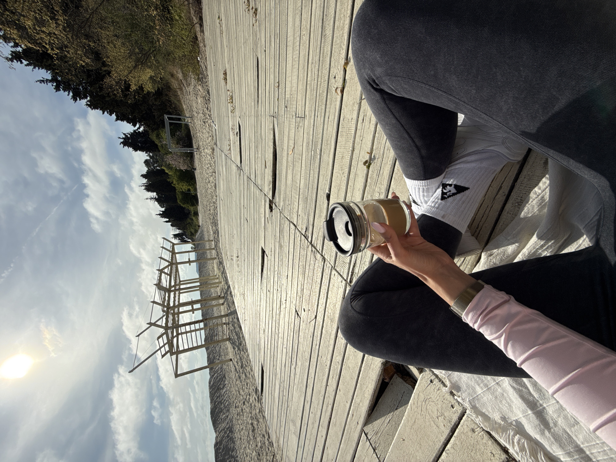 The scene is an eye-level, outdoor shot on what appears to be a wooden boardwalk or deck. A person is sitting cross-legged with their legs visible on the right side of the frame. They are wearing dark grey leggings, white socks with a black design on the ankle, and have a pink long-sleeved shirt or top. Their left hand is holding a glass jar-like container with a black lid, containing a liquid that seems to be coffee.
The boardwalk stretches into the distance, showing the texture and wear of the wooden planks. In the background, to the left, is a beach with sand and water, and there's a man-made wooden structure resembling a viewing platform. Trees line the distant edge of the beach. The sun is visible in the upper left of the frame, suggesting it is daytime.
Based on the location provided (Nesebar, Bulgaria), the scene is likely set along the coast. The interaction is minimal, only showing the person holding a drink, likely enjoying the view or taking a break.