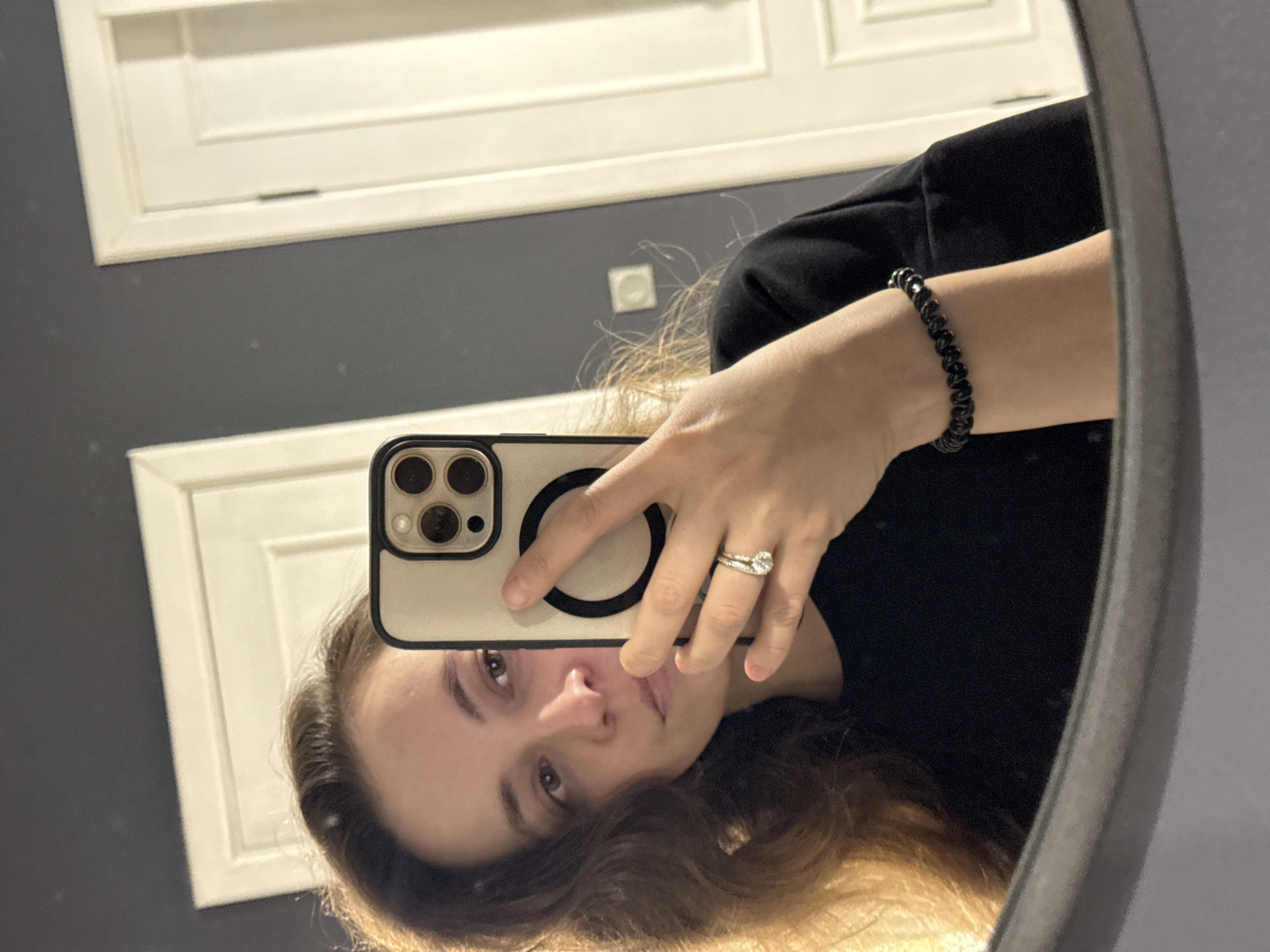 A mirror selfie captures a woman holding a smartphone, reflected in an oval or rectangular mirror. The image is oriented with the woman's head at the bottom, looking up towards the camera. She has long, wavy brown hair. Her left hand holds the phone, which has a light-colored case with a prominent black MagSafe-style magnetic ring. On her left ring finger, she wears two rings, one featuring a round diamond. On her right wrist, she has a black spiral hair tie/bracelet. She is wearing a black top.
The background, visible as a reflection in the mirror, shows a room with dark gray walls. White architectural details, likely door panels or cabinet fronts, are reflected both above and below the woman's figure. An electrical outlet with a white cover is visible on the wall behind her. The woman's expression is neutral as she looks directly at the camera. The lighting suggests an indoor setting. No discernible text is visible.