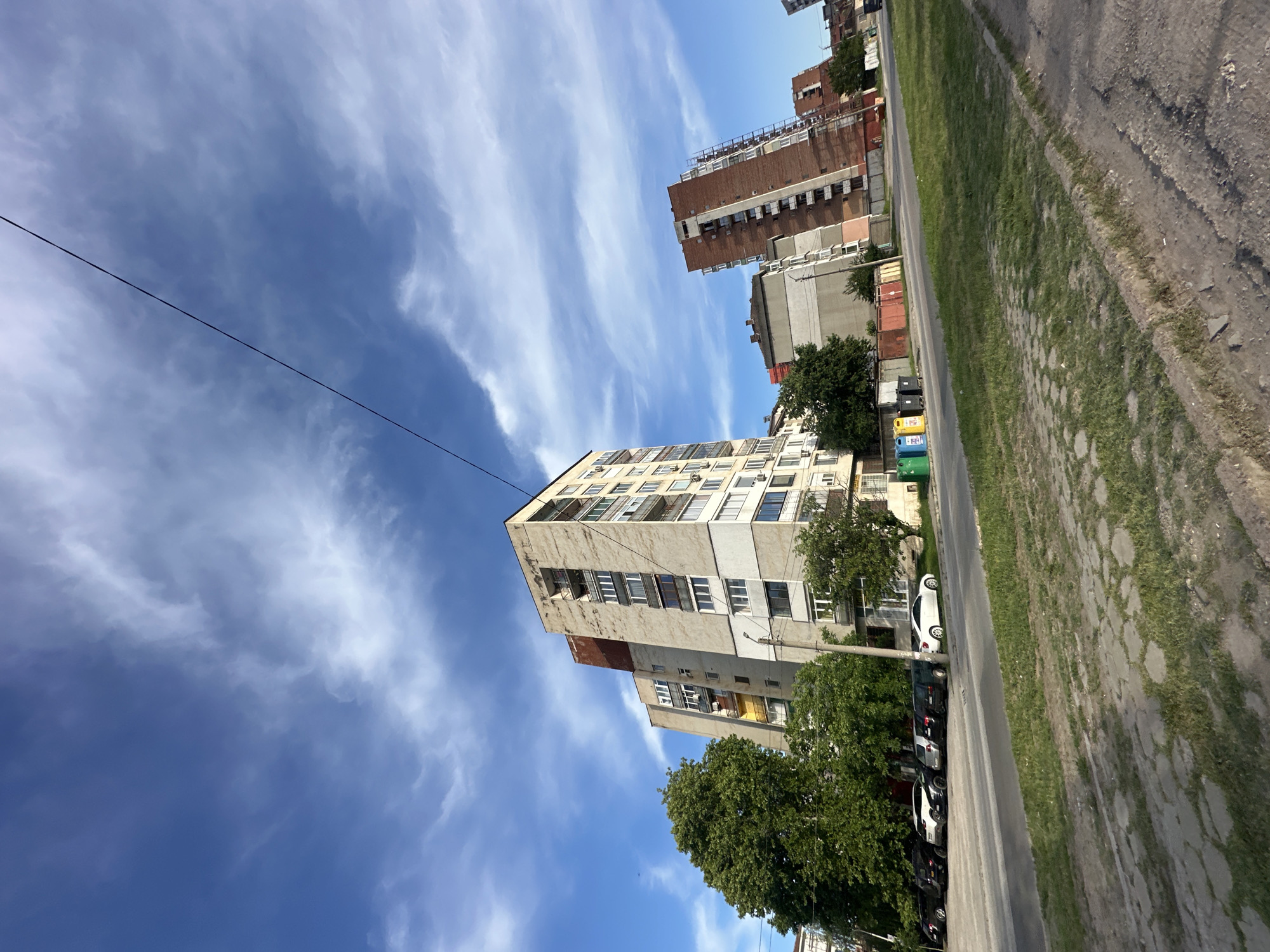 The image depicts an urban scene in Lovech, Bulgaria. The perspective is from ground level, looking upwards at an angle. Prominent features include several multi-story residential buildings, constructed of concrete and brick. The buildings display a mixture of gray and brown facades with windows and balconies. Trees with full green canopies are situated near the buildings, partially obscuring the lower floors. A row of parked cars lines the street, with a variety of colors and models visible. Recycling bins are visible near one of the buildings. A utility wire stretches across the sky. The sky is bright blue with wispy white clouds. The ground surface consists of a paved road with a grassy verge to one side. The overall impression is of a typical residential area in Lovech.