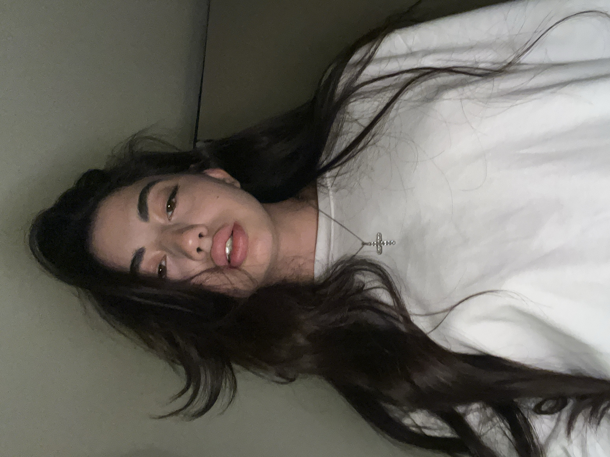 The image shows a medium shot of a young woman looking directly at the camera. She has long, dark, wavy hair that is partially obscuring her face. She is wearing a white crewneck sweatshirt and a silver cross necklace. The background is a plain, dark wall. The lighting is dim, with the light source appearing to be from above and slightly to the front of the subject. The image is taken from a low angle, looking up at the woman. There are no visible indications of the location being Chotěšov, Czechia.