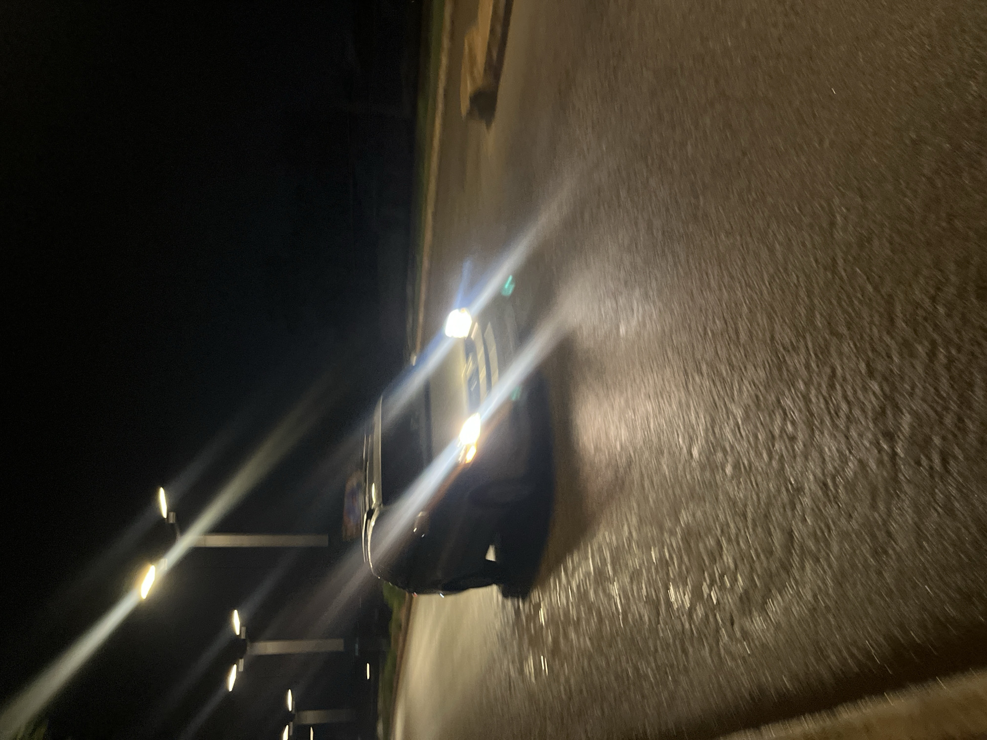 This is a night-time, low-light, motion-blurred photograph taken from a moving vehicle on a paved road in Bukuru, Nigeria. The foreground is dominated by the wet asphalt road, reflecting the bright beams of streetlights and the headlights of a vehicle. In the mid-ground, a dark-colored SUV or van is in motion, its headlights illuminated and casting strong lens flares across the scene. The vehicle appears to be a light-colored SUV or van. In the background, several streetlights with multiple bulbs are visible, lining the side of the road, with their light beams creating streaks across the dark sky. The overall environment suggests an urban or semi-urban street at night, possibly after rainfall, given the wet appearance of the road.