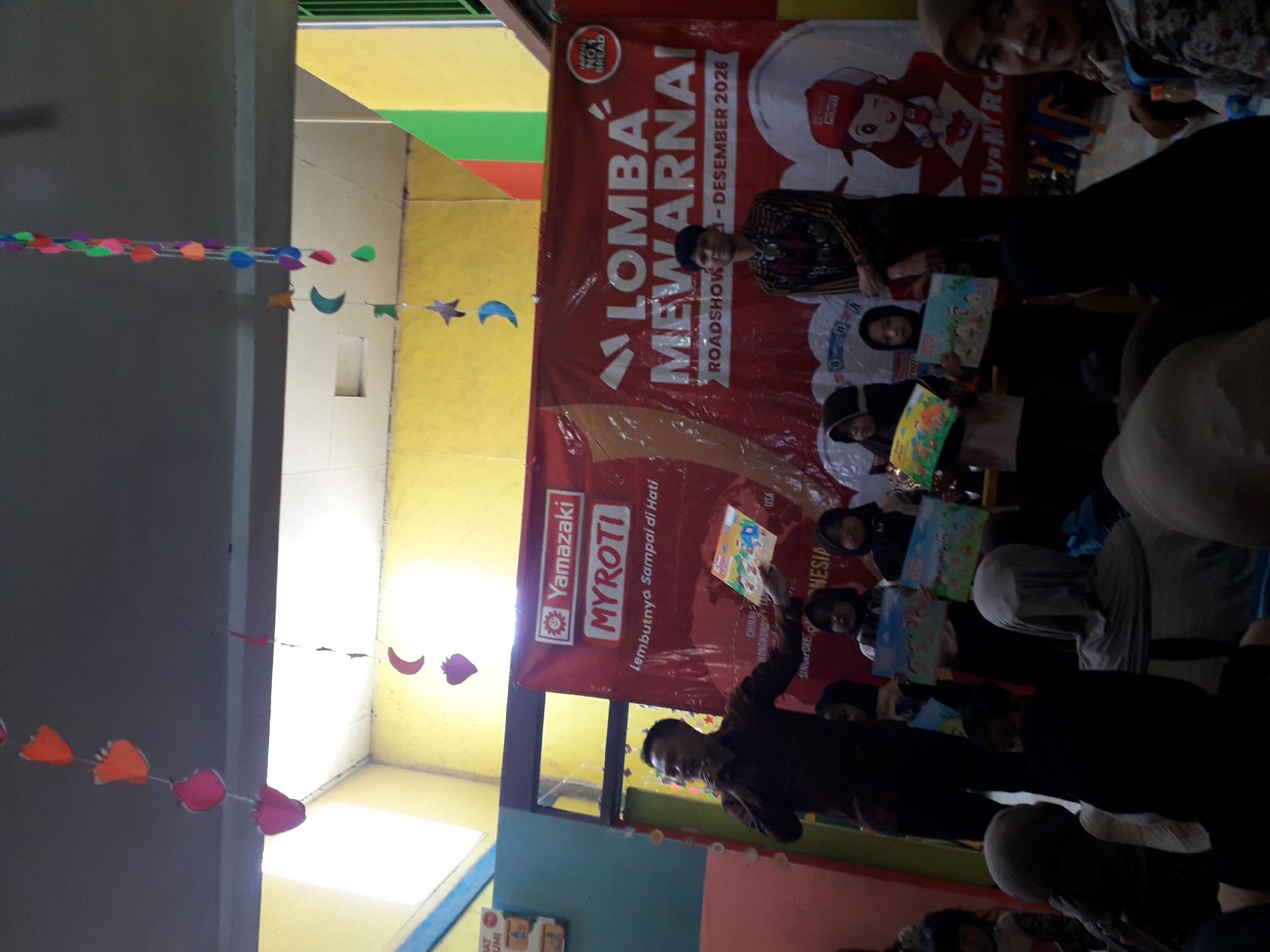 In Cankuang, Indonesia, an event, possibly a children's drawing competition, is underway indoors. A banner behind a group of people reads "LOMBA MEWARNAI ROADSHOW 21 - DESEMBER 2026," indicating a coloring competition roadshow. A man wearing a dark long-sleeved shirt is holding a microphone and a drawing, speaking to an audience. To his right, another man in traditional attire stands beside the banner. In front of them, several children, mostly girls wearing hijabs, are holding drawings. One child is holding a trophy. Decorative paper cutouts of fruits, moons, and stars hang from the ceiling, suggesting a festive atmosphere. The walls are painted yellow and green. The overall scene captures a community event focused on children's creative activities.
