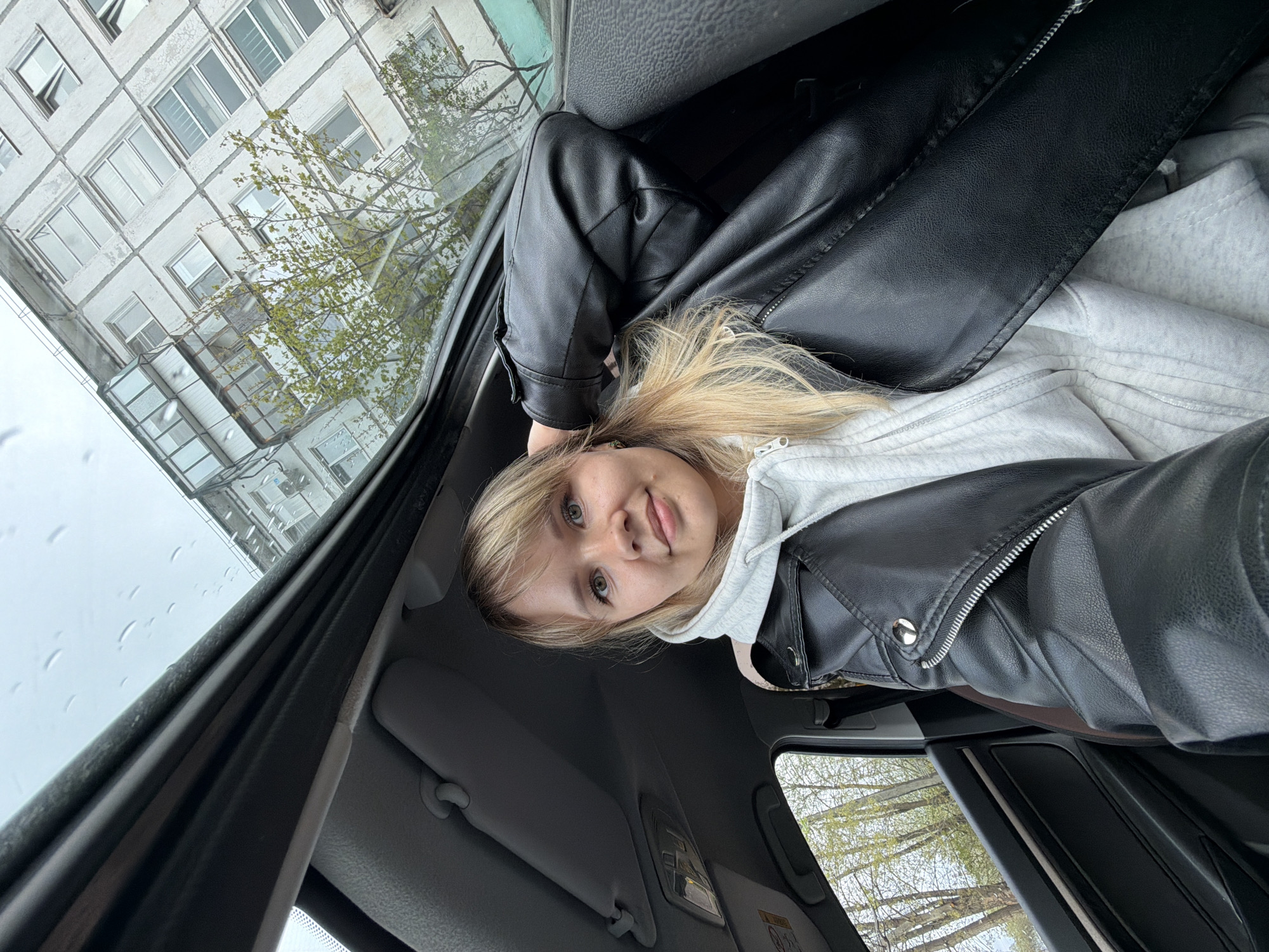 The image is taken from inside a car, looking up at a young woman in a black leather jacket over a light grey hoodie. The woman has blonde hair and fair skin. Through the car's sunroof, a grey, multi-story building with numerous rectangular windows is visible. Bare tree branches are also visible outside the car. Raindrops are present on the sunroof. Based on the location tag, this scene is likely in Chaikovsky, Russia.