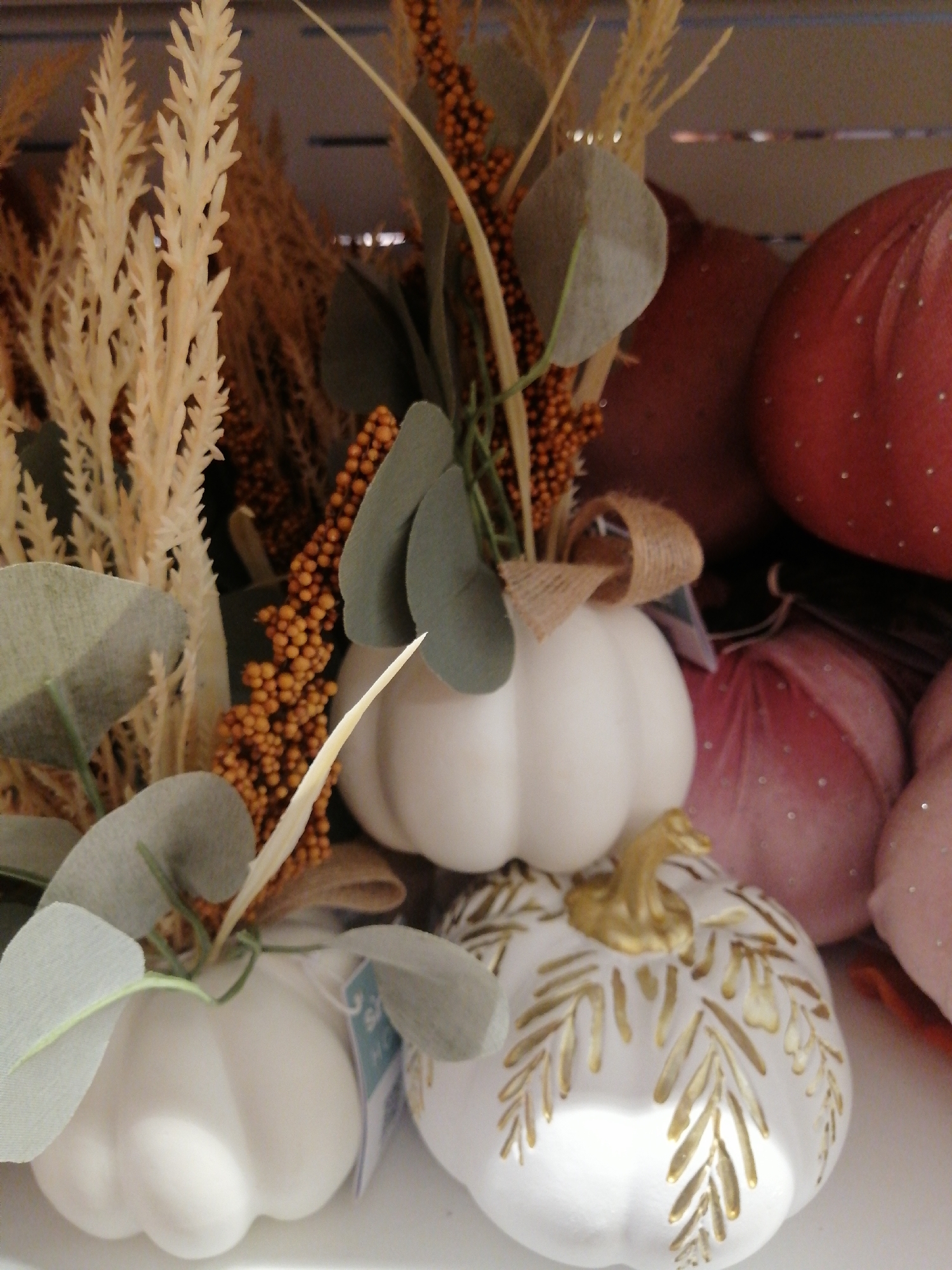 The image displays a close-up of several decorative pumpkins and autumnal foliage. In the foreground, two white pumpkins are visible. The pumpkin on the left is partially obscured by green leaves and a straw-like decorative element. The pumpkin on the right is more prominently displayed, with gold painted leaf designs and a golden stem. Behind these, a cluster of orange berry-like decorations and dried grasses are arranged. In the background, there are larger, rounded decorative items that resemble pumpkins, one deep red and others in shades of pink, all adorned with small, sparkling dots. The overall scene suggests a display of seasonal decorations, possibly in a store or home environment. There are no people or specific city landmarks visible, therefore, the location in Kremenchuk, Ukraine cannot be determined from the image.