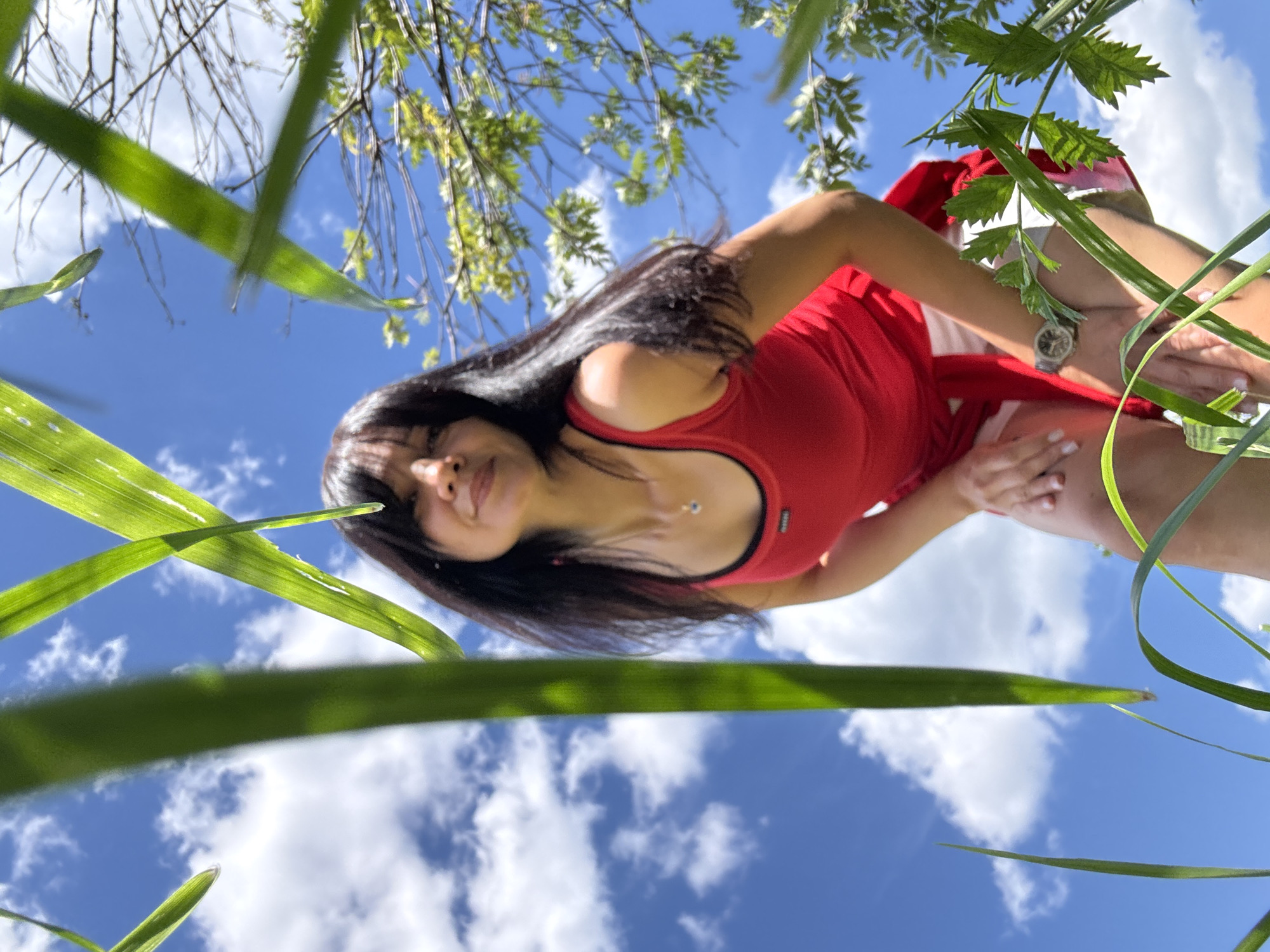 A low-angle, eye-level shot captures a woman with long, dark hair and a red tank top, crouching down in an outdoor environment. She is partially obscured by tall green grass in the foreground, which frames the scene. The woman is looking towards the camera with a slight smile. Behind her, a bright blue sky with scattered white clouds and green trees suggests a sunny day. Her left arm is bent, with her hand resting on her left thigh, and her right hand is extended towards her right thigh. She is wearing white shorts and a watch on her left wrist. The lighting indicates it's daytime, with sunlight illuminating the woman and the surrounding vegetation. The image appears to be taken in a natural setting, possibly a park or a garden. Without additional information, it is not possible to definitively determine if the image was taken in the city of Chaikovsky, Russia, or any specific location.