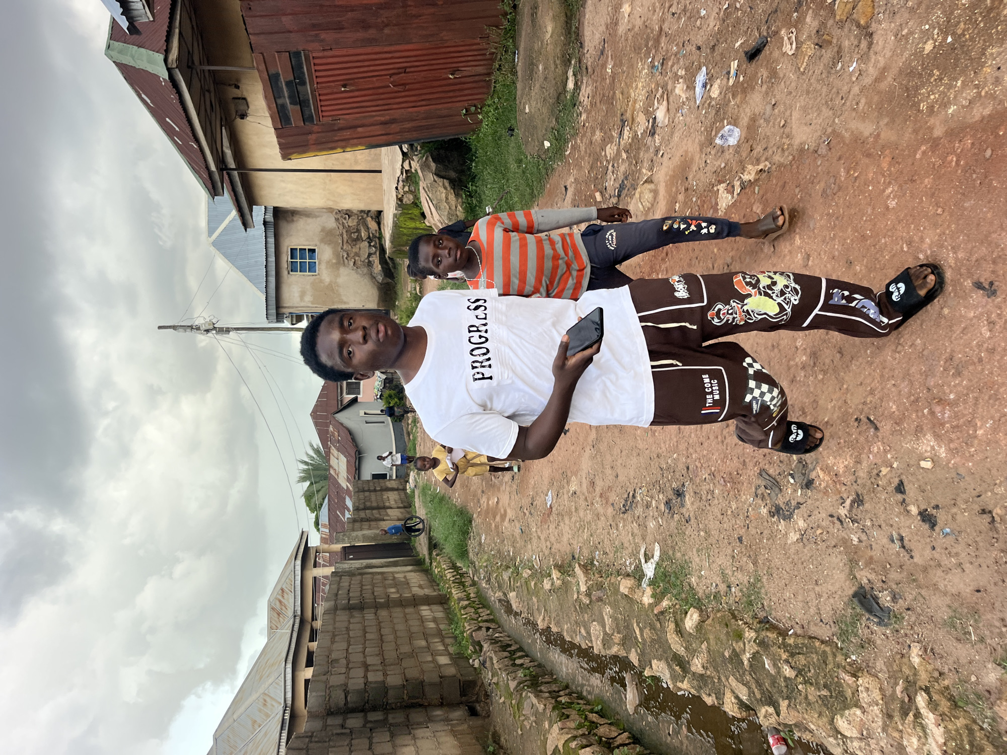 This media file captures an outdoor scene with several people and a dirt path. The primary focus is on two young Black men standing in the foreground. The man on the left is wearing a white t-shirt with the word "PROGRESS" printed on it and dark pants with colorful graphics. He is holding a smartphone in his right hand. The man on the right is wearing a striped long-sleeved shirt in orange and gray, and dark pants. Behind them, several other individuals, including children, are visible in the background, engaged in various activities.
The setting appears to be a somewhat impoverished urban or semi-urban area, characterized by unpaved ground, weathered buildings with corrugated metal roofs, and a drainage ditch running alongside the path. There are scattered pieces of debris and vegetation on the ground. The sky is overcast with heavy clouds, suggesting a potentially gloomy or upcoming rainy weather. The overall atmosphere is casual and appears to be daytime, though the exact time is not discernible. The expressions on the faces of the main subjects are neutral.
Visible text includes "PROGRESS" on the white t-shirt of the man on the left. On the pants of the man on the left, there is text that appears to read "THE COME MUSIC".