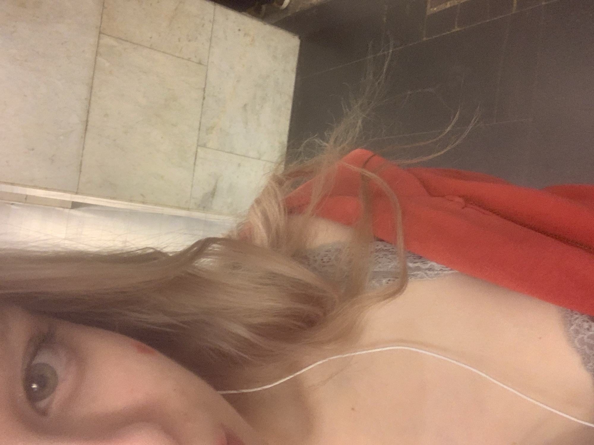 A close-up, rotated image shows a person from a low angle, possibly lying down, with their left eye, nose, and cheek visible on the left side. The person has long, flowing blonde or light brown hair that extends across the frame and partially obscures their shoulder and upper back. A thin white cord, likely from headphones, crosses their chest area. They are wearing a red garment, possibly a jacket or hoodie, on the right side of the image, and a light-colored, possibly lace-patterned undergarment is visible beneath it. The background consists of two distinct areas: the upper portion shows light-colored, square-tiled flooring, resembling marble or ceramic, while the lower portion features a darker, plain surface, possibly another type of flooring or a wall. The lighting appears to be artificial and somewhat dim. There is no visible text in the image. Given the context, this scene is located in Saint Petersburg, Russia, though no specific visual cues within the image confirm this.