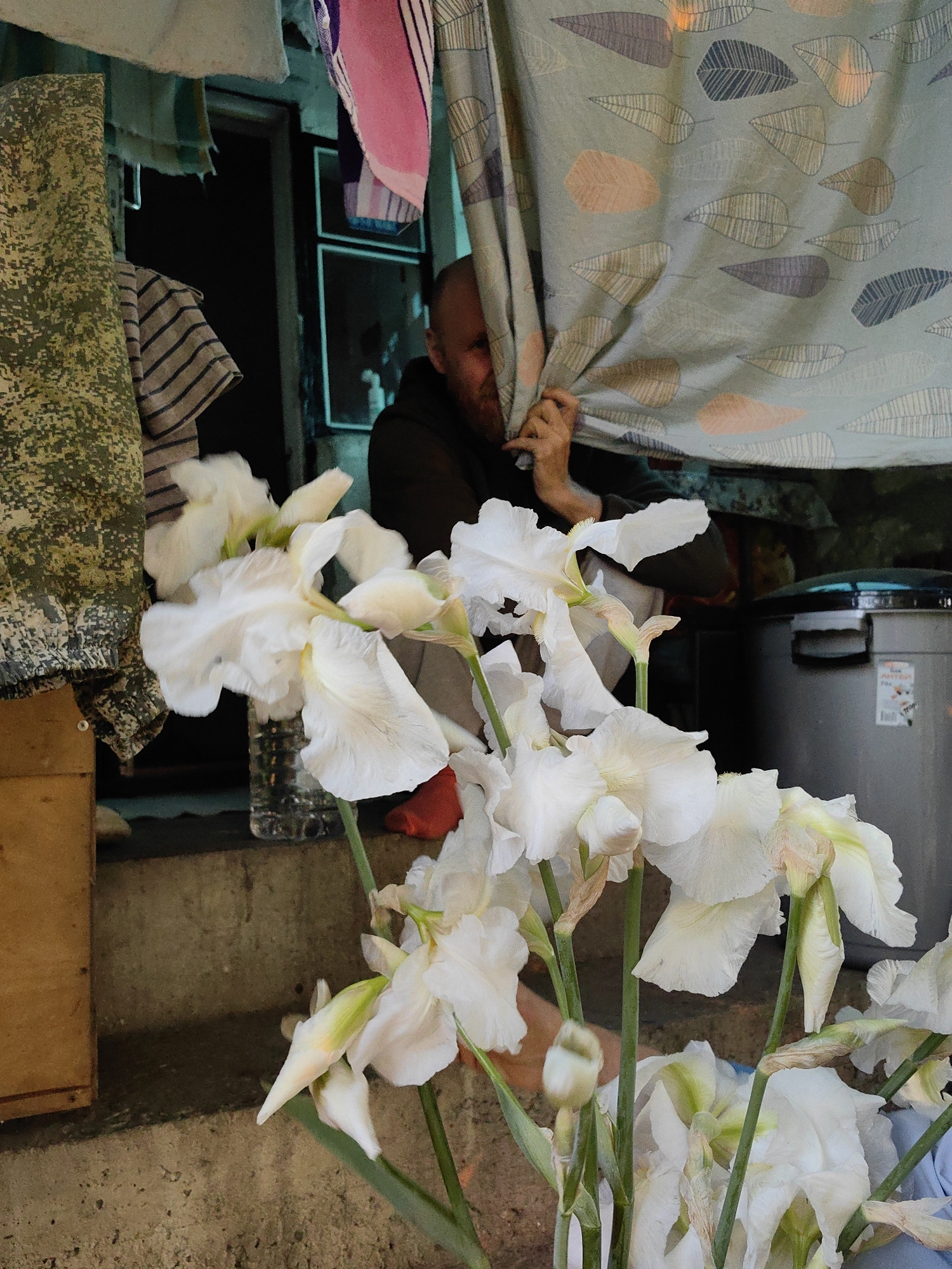A man partially obscured by a patterned curtain looks out from a doorway crowded with clothing and household items in what appears to be a residence in Зуйское сельское поселение, Ukraine. A large bouquet of white irises dominates the foreground. The setting appears cluttered and improvised.