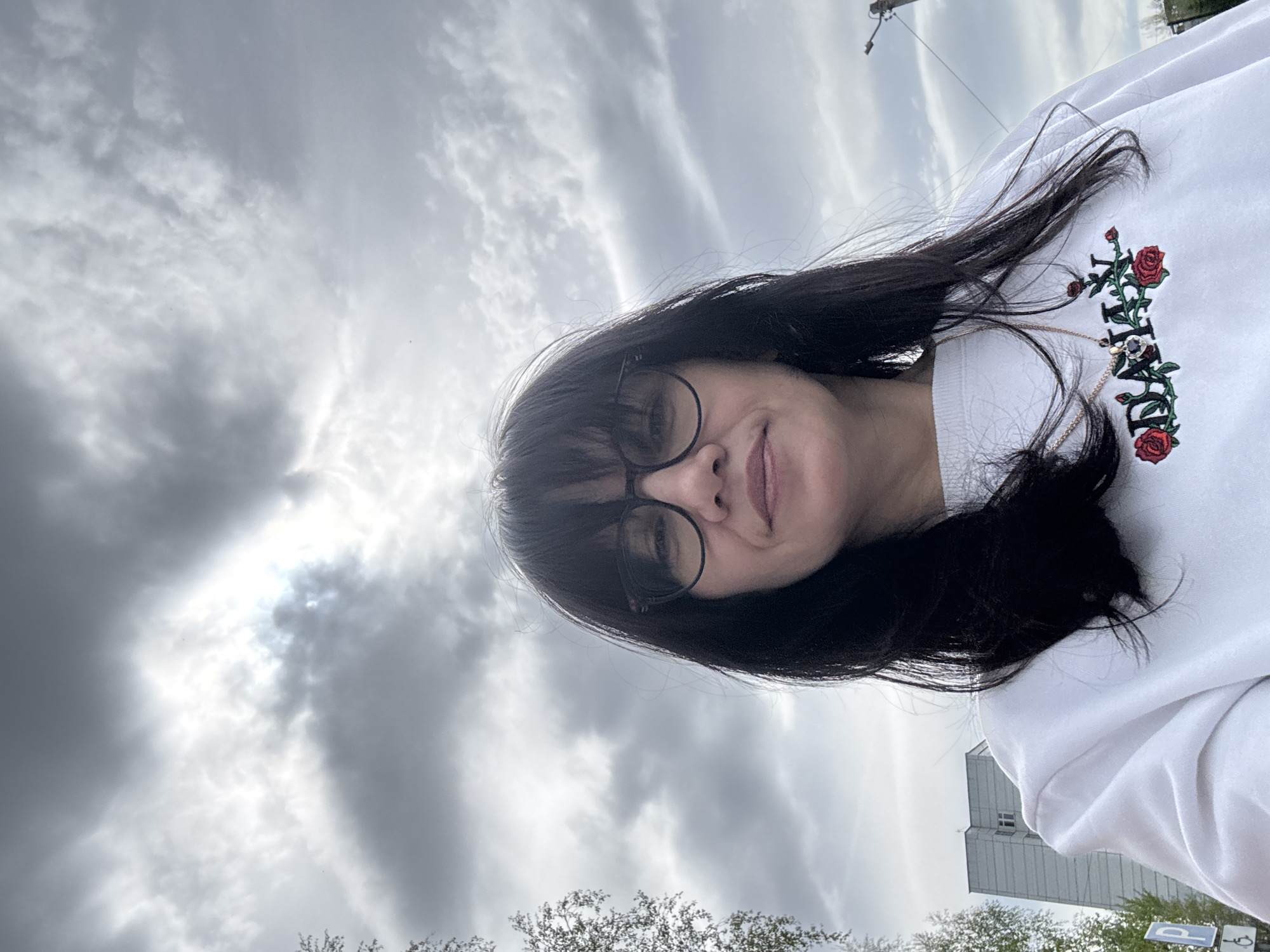 A woman with dark hair and glasses is posing for a selfie in Chaikovsky, Russia. She is wearing a white sweatshirt with an embroidered floral design. Behind her, the sky is overcast with dramatic clouds. A building can be seen in the distance, and some trees are visible at the bottom of the frame. Overhead power lines are also visible. The location appears to be outdoors, potentially near a park or green space in Chaikovsky.