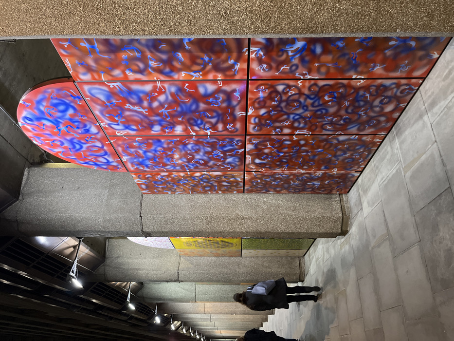 An unusual perspective presents a subterranean space with concrete structures overhead, adorned with red and blue abstract art composed of stylized figures. People descend tiled stairs in the foreground. Visible are air vents with illuminated lights beneath a bridge, partially revealing colorful murals on the ceiling, creating a sense of depth and dimension.