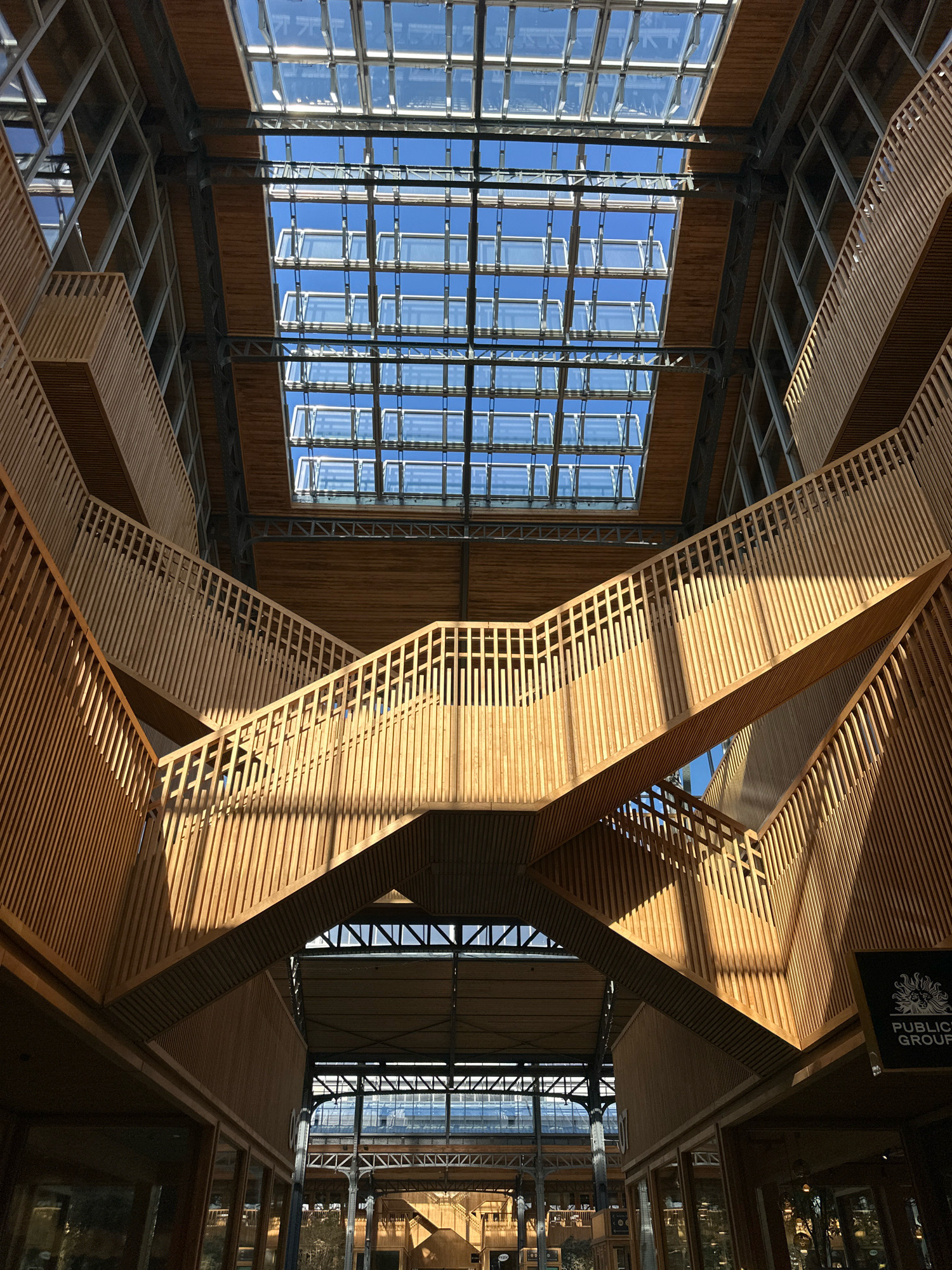 The image shows the interior of a building, focusing on a complex wooden staircase structure. The staircase has numerous levels and is made of light-colored wood with vertical slats forming the balustrades. Sunlight streams through a large glass roof, casting strong shadows across the wooden surfaces. The architecture appears modern and geometric, with a strong emphasis on natural light and materials. In the lower right corner, a black sign with a circular emblem and text that reads "PUBLIC GROUP" is visible. The building's interior also shows exposed metal structural elements and wooden paneling on the walls and ceilings. The overall impression is one of an open, airy, and architecturally striking public space. The location is the Public Group building in Molenbeek-Saint-Jean - Sint-Jans-Molenbeek, Belgium.