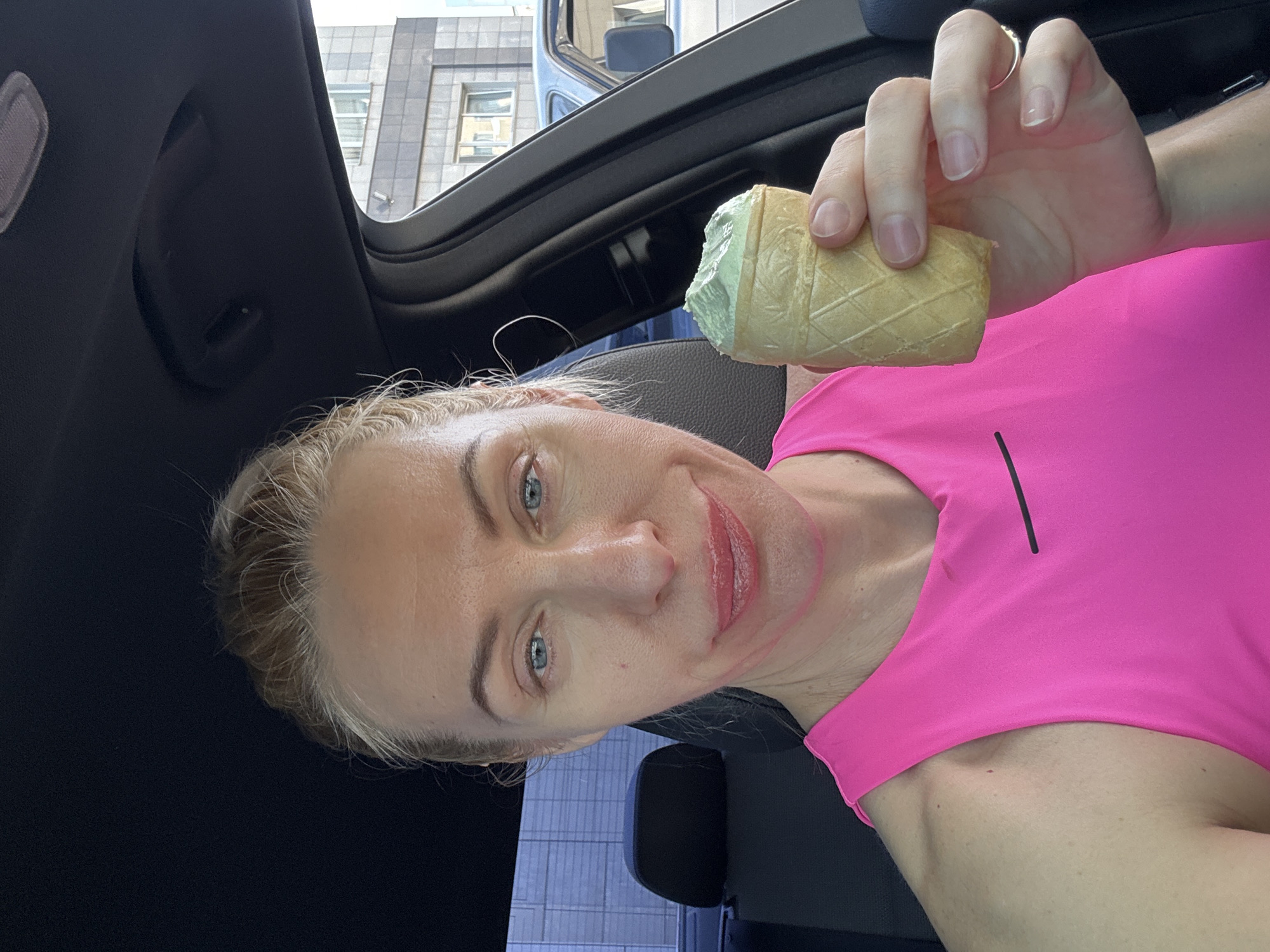 A woman is sitting in a car while holding a green ice cream cone. She is wearing a bright pink athletic shirt. The car is parked in a city, with a tall building visible through the window.