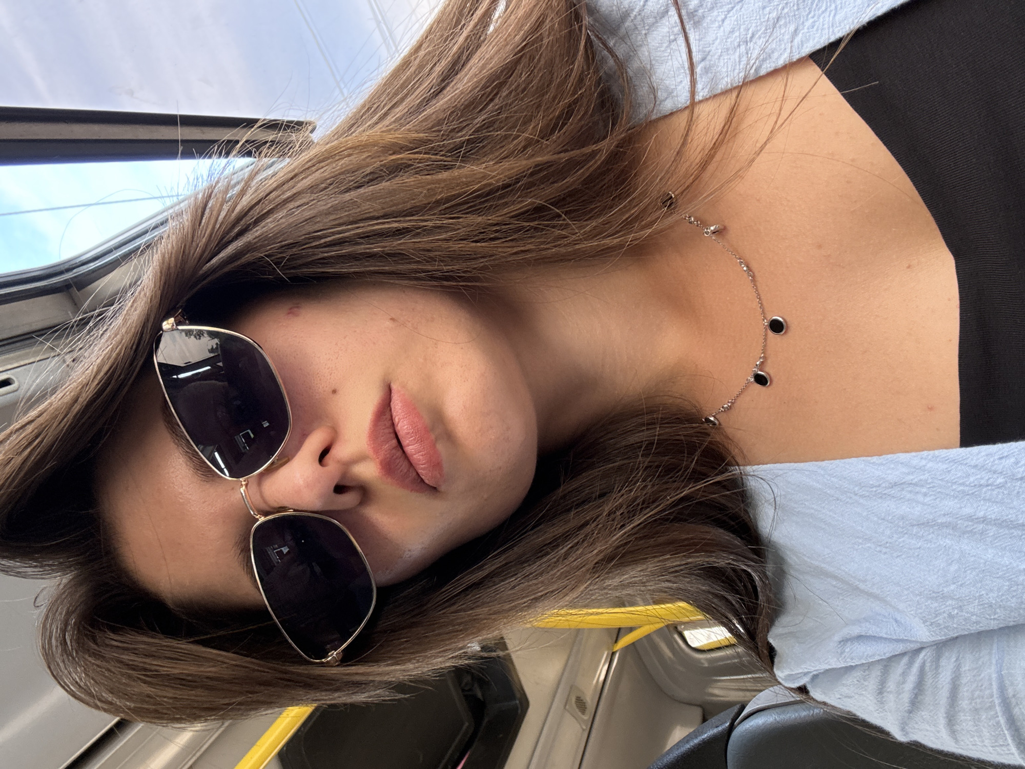 A medium shot captures a young woman with long, brown hair and sunglasses, facing the camera. She is indoors, appearing to be in a bus or train, with yellow poles visible in the background. She is wearing a light blue, textured jacket over a black top and a delicate silver necklace with three dark circular pendants. The lighting suggests daytime.