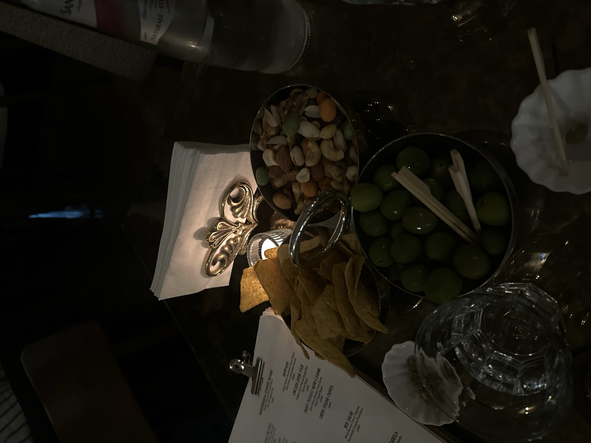 The image depicts a table setting, likely in Yerevan, Armenia. Present are a bottle of "SAN NATURALE" water, a bowl of mixed nuts, a bowl of green olives with toothpicks, tortilla chips, a napkin holder with decorative detail, a small white dish, a patterned glass, and a menu on a clipboard. The scene is dimly lit, suggesting an evening setting. The menu appears to list items such as "CHICKEN LIVER PATE" and "GREEN ITALIAN OLIVES."