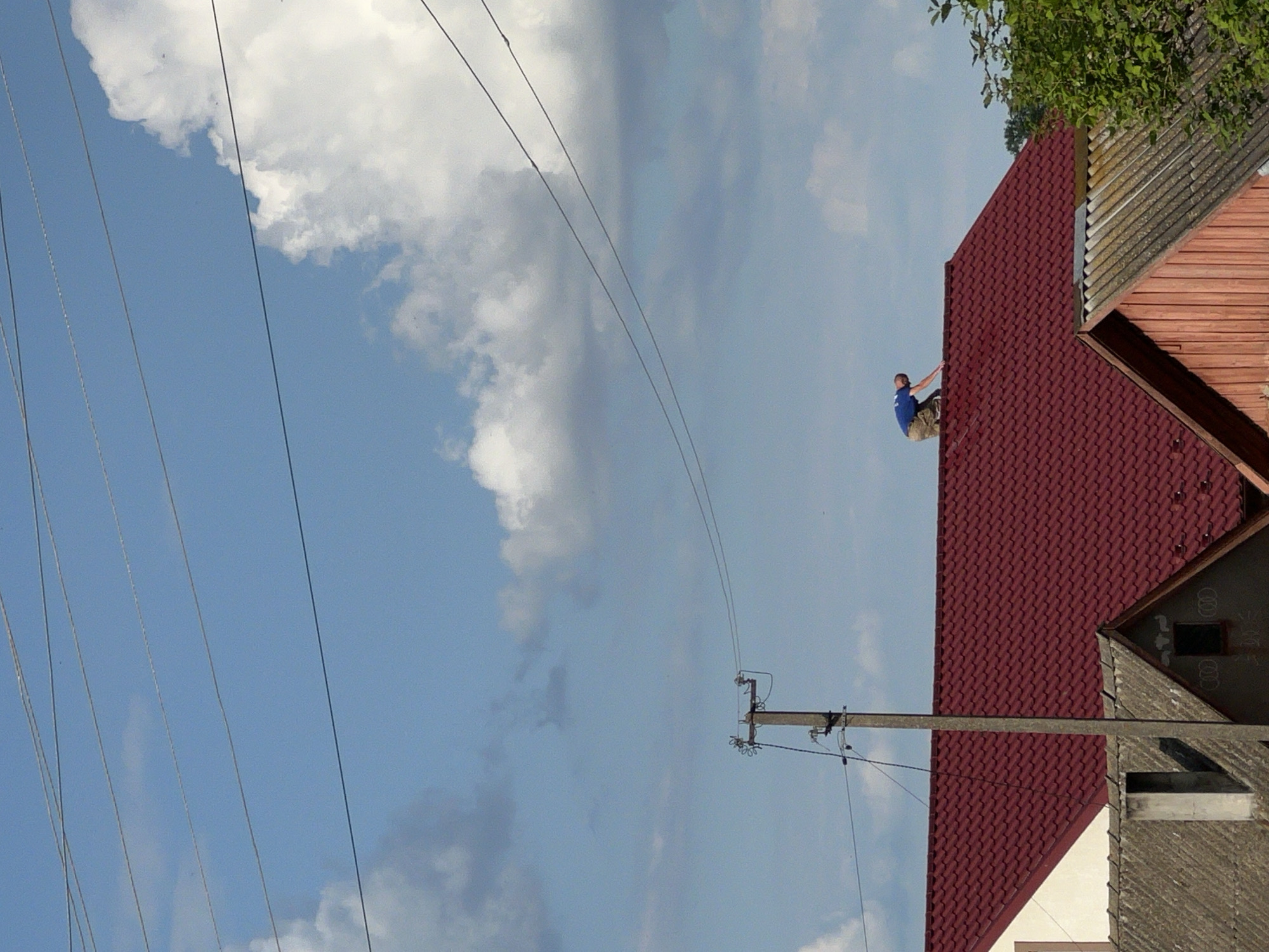 In the city of Vertelka, Ukraine, a man in a blue t-shirt and camouflage shorts is on his hands and knees on the bright red, tiled roof of a house. He appears to be working on the roof. The house has a white gable on the left side. In front of this house, another house with a grey, tiled roof and a chimney is visible. To the right of the red-roofed house, a tree with green leaves is present. A utility pole with wires stretches across the scene, with multiple wires also visible against a blue sky dotted with white clouds. The image is taken from a low angle, looking up at the houses and sky.