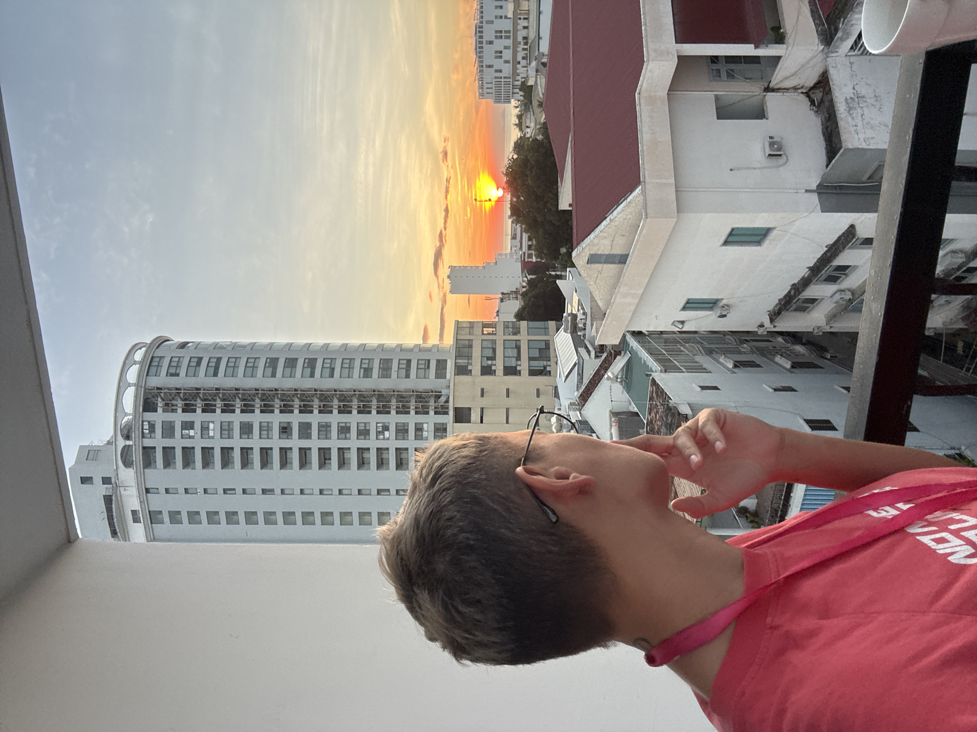 A person with short, blond hair and glasses stands on a balcony, looking out at a cityscape during sunset. The person is wearing a pink t-shirt and a pink lanyard. In the background, several buildings of varying heights are visible, with a tall, light-colored tower dominating the left side of the frame. The sun is low on the horizon, casting a warm orange glow across the sky and illuminating some of the buildings. The scene appears to be in Nha Trang, Vietnam, a coastal city known for its beaches and high-rise hotels. The architecture suggests an urban environment.