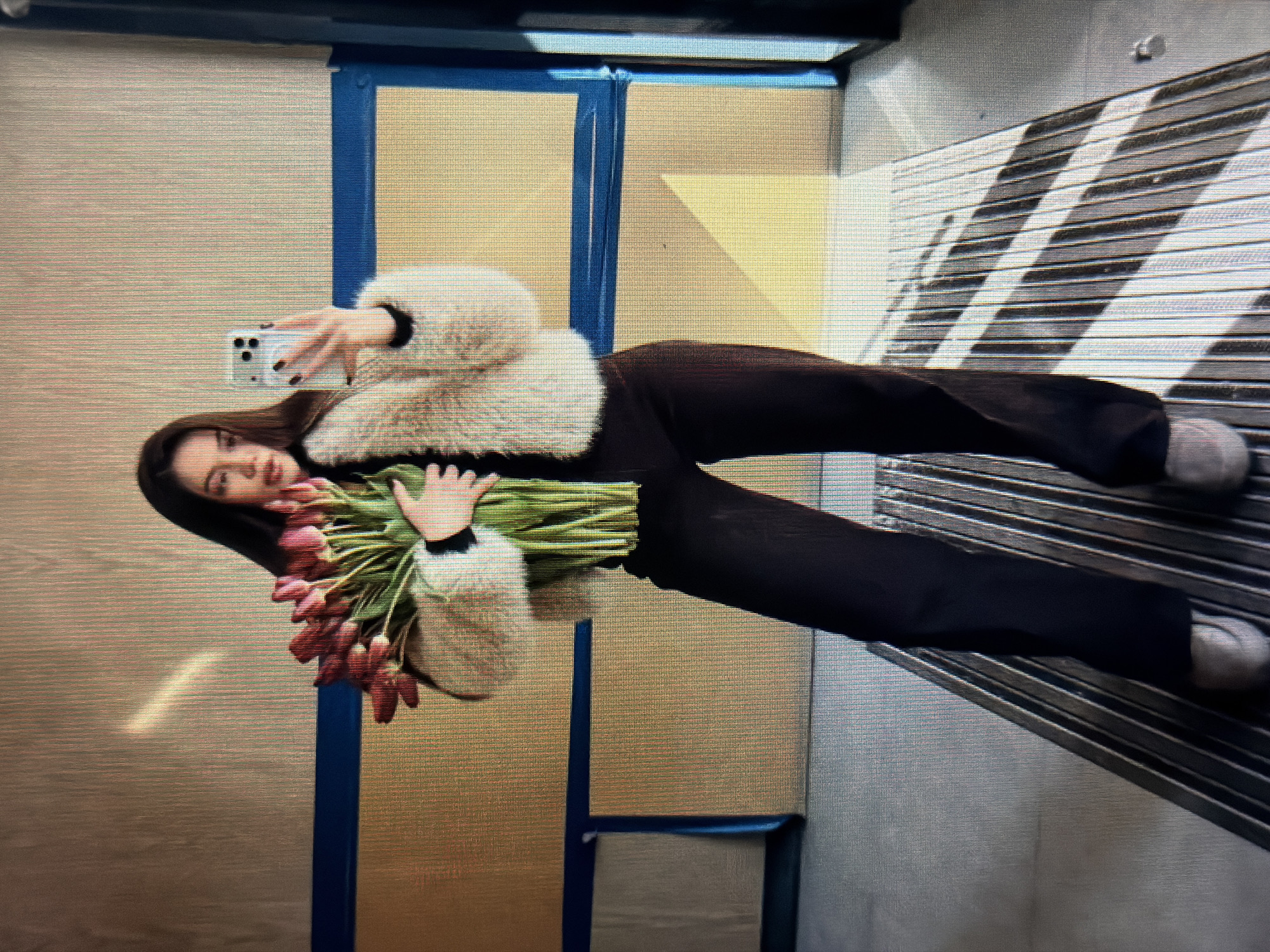 A young woman with dark hair is standing indoors, holding a large bouquet of tulips. She is wearing a fluffy, light-colored jacket over a black top and dark pants. She is taking a selfie with a smartphone held in her right hand. The background appears to be a wall with horizontal blue and tan stripes. The floor is metallic and slatted, with shadows cast across it, suggesting the presence of a light source from the side. The overall lighting is bright, with some sunbeams visible. The location is identified as the city of Sirius, Russia.