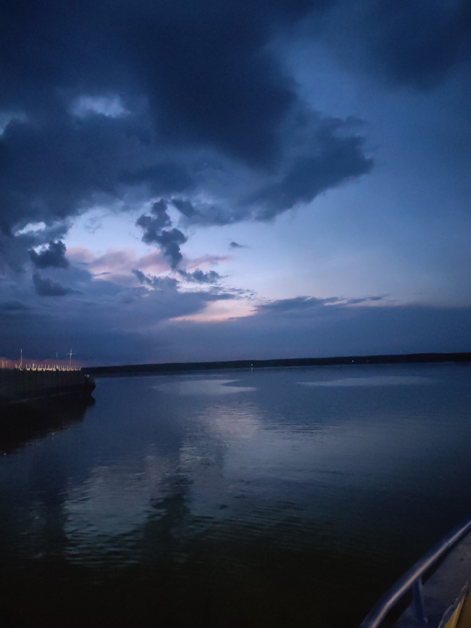 The image depicts a twilight scene over a body of water, likely a river, with a dramatic sky filled with dark, heavy clouds tinged with soft hues of pink and pale blue from the setting sun. In the foreground, the dark water reflects the moody sky. To the left, a portion of a large vessel or structure is visible, featuring a row of lights along its edge. This structure appears to be a pier or a large boat docked alongside. The water is calm, with subtle ripples indicating a gentle breeze. The overall mood is tranquil yet imposing, characteristic of the transition from day to night under a stormy or overcast sky. The location is identified as Rybinsk, Russia. No visible text is present in the image.