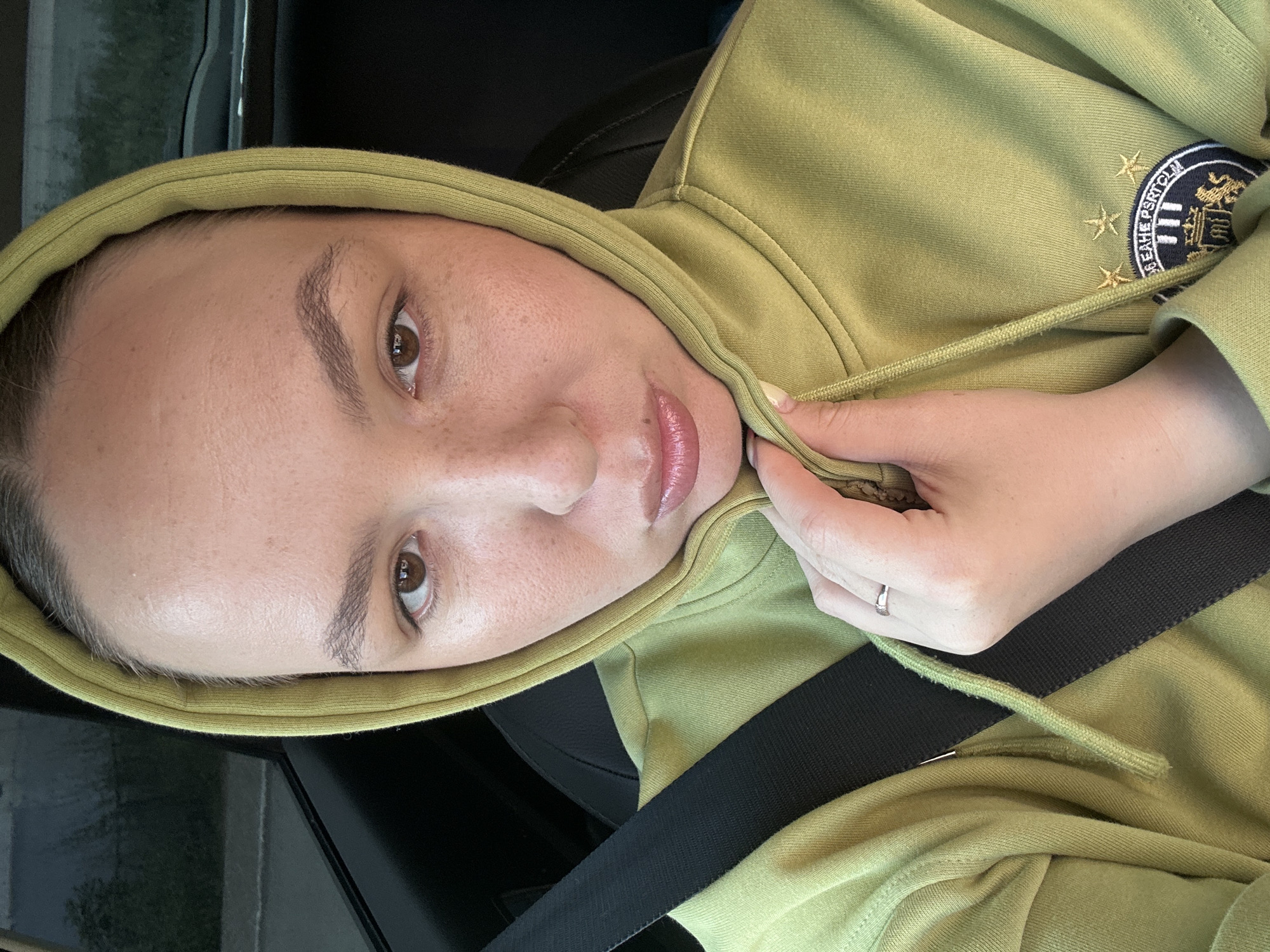 A medium shot photo captures a woman wearing a light green hooded sweatshirt. She is seated in what appears to be a car, with a black seatbelt visible across her chest. The woman is looking directly at the camera, with her lips slightly pursed. Her hand is holding the hood of her sweatshirt near her chin. A ring is visible on her finger. There is an emblem on the lower right side of the sweatshirt. The background is out of focus, suggesting the car is in motion or parked. This image was taken in the city of Chaikovsky, Russia.