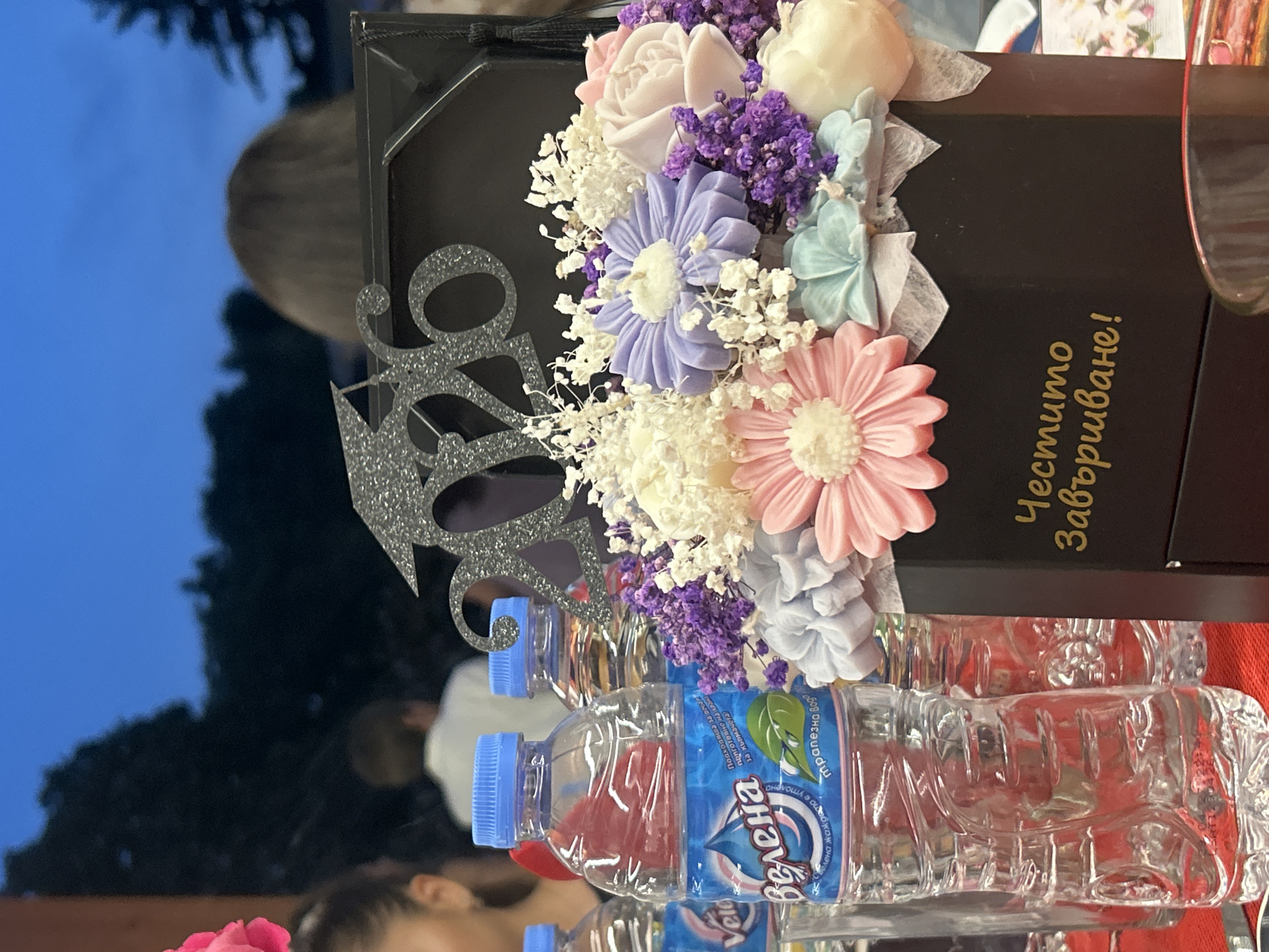 The image captures a detailed view of a graduation celebration, specified to be in Kaspichan, Bulgaria. In the immediate foreground, a black hexagonal box serves as a centerpiece, containing an arrangement of artificial flowers. These flowers are varied in color, including light pink, lavender-purple, cream-white, and light blue/teal, and are interspersed with white baby's breath, suggesting a decorative bouquet, possibly composed of soap or wax. A silver glittery cutout displaying the numbers "2026" and a graduation cap sits prominently at the back of the box, indicating the year of graduation. Gold Cyrillic text, "Честито завършване!", meaning "Happy Graduation!", is embossed on the front face of the box.
To the left of the centerpiece, multiple clear plastic water bottles with bright blue caps are visible. The label of the closest bottle clearly shows the brand name "Велена" (Velena) and "трапезна вода" (table water), accompanied by a logo featuring a water droplet and a green leaf. These bottles rest on a red surface, likely a tablecloth. A segment of a clear drinking glass is visible in the bottom right corner of the frame.
In the midground, several individuals are partially visible behind the table setup. On the far left, a portion of a person's head with dark hair is seen. Further back, the upper torso and back of a person wearing a white shirt are visible, facing away from the camera. To the right of the center, another individual's back and brown hair are discernible, also facing away. The background consists of a dark blue sky, characteristic of dusk or nighttime, with the indistinct silhouettes of trees and dense foliage against it, suggesting an outdoor or semi-outdoor setting for the event.