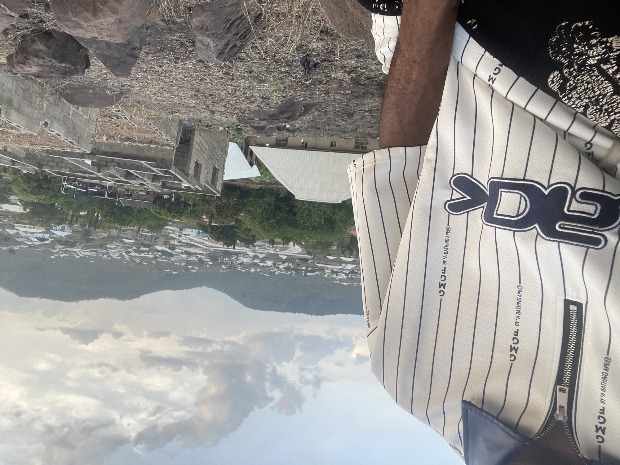 The image, if rotated to landscape orientation, captures an outdoor scene in Lokoja, Nigeria, from an elevated perspective. A person is partially visible in the foreground on the right, showcasing a dark-skinned arm and torso. They are wearing a white pinstriped shirt or jacket. Notable text visible on the garment includes "BY A BATHING APE®" repeated several times, "GMGF" (or "CMGF" as per OCR), and a large, stylized black logo that appears to be "BAPE". A metal zipper is also visible on the sleeve of the garment.
The immediate foreground consists of dry, rocky ground with sparse, light-colored vegetation. Large, dark boulders are scattered across this area. Behind this, there are structures under construction, built with concrete blocks and exposed rebar, indicating ongoing development. Further into the midground, a dense urban or semi-urban area spreads out, characterized by numerous residential buildings with varying roof designs, interspersed with greenery from trees. In the background, prominent, undulating hills or mountains rise, bordering the settlement. The sky is overcast with a mix of grey and lighter clouds, suggesting a cloudy daytime scene. The image provides a glimpse of a developing town set against a natural, hilly landscape.