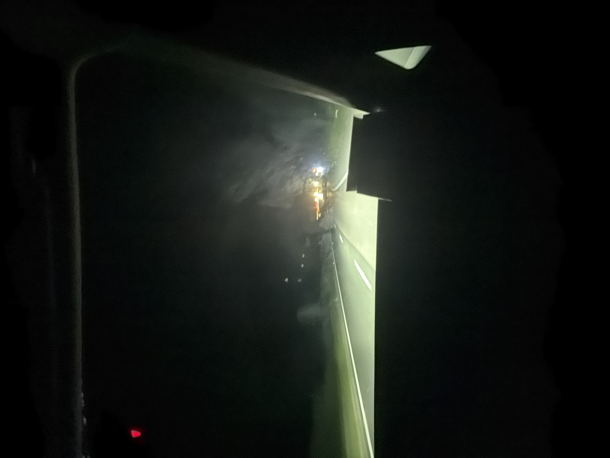 The image depicts a nighttime view from inside a vehicle, looking out through the windshield. The road ahead is illuminated by headlights, with white lines marking the lanes. In the distance, there are clusters of bright lights, suggesting a settlement or buildings. The surroundings are dark, with faint outlines of trees or landscape. A small red light is visible on the top left of the windshield. The scene appears to be on a road leading towards the city of Razliv, Bulgaria, likely during a drive.