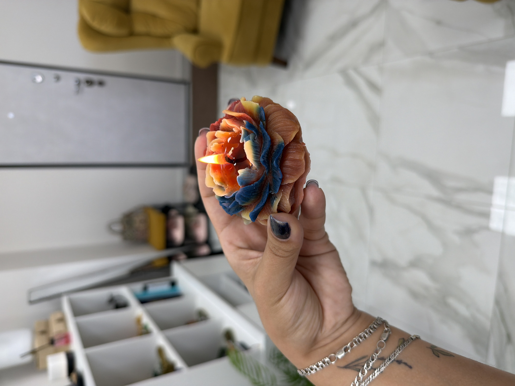A hand with dark nail polish and silver bracelets holds a lit, multi-colored flower-shaped candle. The candle has petals in shades of orange, yellow, and blue, with a flame burning in the center. The background is blurred, showing a white shelf unit with items on it, a grey door, and a yellow armchair. The floor appears to be a marble-patterned tile. This scene is located in a room, not a specific outdoor city location.
