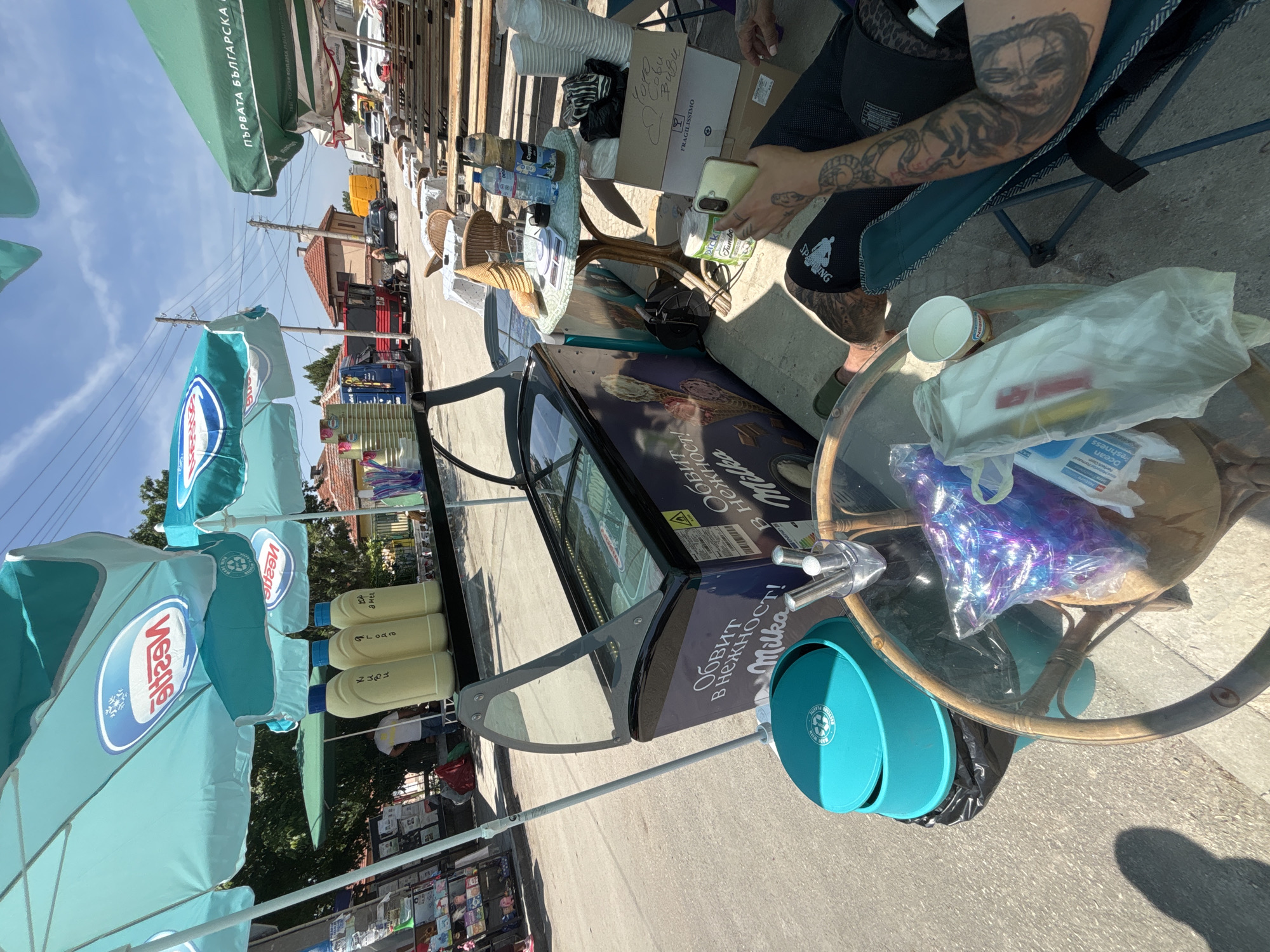 The scene depicts an outdoor vendor setup in Kostievo, Bulgaria, on a clear, sunny day. Multiple umbrellas provide shade; three prominent turquoise umbrellas feature the inverted red and white "Nestlé" logo, viewed from below, and a large green umbrella on the right displays Bulgarian text reading "ПЪРВАТА БЪЛГАРСКА" (The First Bulgarian).
In the center, a dark blue commercial ice cream freezer or display case is visible, with one glass lid partially open. The freezer is branded "Milka" and features the Bulgarian phrase "ОБИТ В НЕЖНОСТ!" (Wrapped in tenderness!). Behind the freezer, on a black stand, three yellow plastic squeeze bottles with blue caps are arranged, bearing handwritten Cyrillic labels. Two are identifiable as "киви" (kiwi) and "ягода" (strawberry), with the third partially visible.
In the immediate foreground, a small, round, glass-topped table with a wicker-like base holds a transparent plastic bag containing shiny purple items, a white plastic bag, and a light blue paper cup with a Nestlé logo. To the left of this table, a turquoise trash bin with a recycle symbol is present. To the right of the ice cream freezer, another small, round table with a patterned white and blue cloth supports a digital scale, two plastic water bottles (one labeled "Cappy"), and a stack of ice cream cones. A stack of white paper cups is on the ground next to this table, along with a cardboard box marked "FRAGILISSIMO" and handwritten Cyrillic text. A clear container holds several metal spoons or scoops.
A person is seated in a dark blue patterned folding chair on the right side of the frame, holding a light-colored smartphone in their right hand. Their right forearm is heavily tattooed with dark, intricate designs. Their legs are partially visible, wearing dark clothing and a light-colored sandal on the left foot. Another person's legs are partially visible further left in a green sandal. In the background, a person wearing a white t-shirt and blue jeans stands near a blue van.
The background shows a street or open area with various vehicles, including a blue van, a red truck, a yellow truck, and several cars parked in the distance. Buildings with traditional reddish-brown tiled roofs are visible behind the vehicles, and overhead power lines crisscross the bright blue sky. The ground is a mixture of asphalt and lighter concrete or pavement, with distinct shadows indicating strong sunlight.