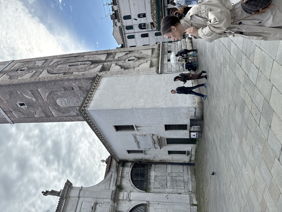 The image captures a daytime scene in Campo Santo Stefano, Venice, Italy, featuring the leaning Campanile di Santo Stefano and a portion of the Chiesa di Santo Stefano. In the immediate foreground, a young adult female is seen in profile, facing right, from the chest up. She wears a light-colored trench coat and carries a dark brown circular patterned bag with a light strap over her left shoulder, with her right hand near her collarbone.
The midground is dominated by architectural elements. The Campanile di Santo Stefano, a tall, light-grey brick bell tower with distinct decorative patterns and rectangular and arched openings, rises diagonally from the center-bottom towards the upper right, exhibiting a visible lean. To the tower's left is a smaller, two-story white-plastered building featuring a dark green entrance door, barred windows, and an ornate rectangular plaque positioned above the door. Adjacent to this white building, on the far left, is a section of the larger, grey stone facade of the Chiesa di Santo Stefano, characterized by large arched windows covered with grates and a statue of a robed figure atop its upper edge.
On the grey stone-paved square in the midground, two individuals walk from left to right: a man in a dark jacket, blue jeans, and blue shoes, followed by a woman in dark attire looking at a smartphone. Further back on the right, beneath a white umbrella, an individual is seated at a small table, with another person standing nearby, indicating an outdoor vendor or artist setup. Two dark grey waste bins are positioned near the entrance of the white building. A single pigeon walks on the paving stones in the lower-left quadrant. Traditional Venetian buildings with tiled roofs and windows are visible in the background on the right. The sky is largely overcast with white and grey clouds, though a patch of clear blue sky is present in the upper right.