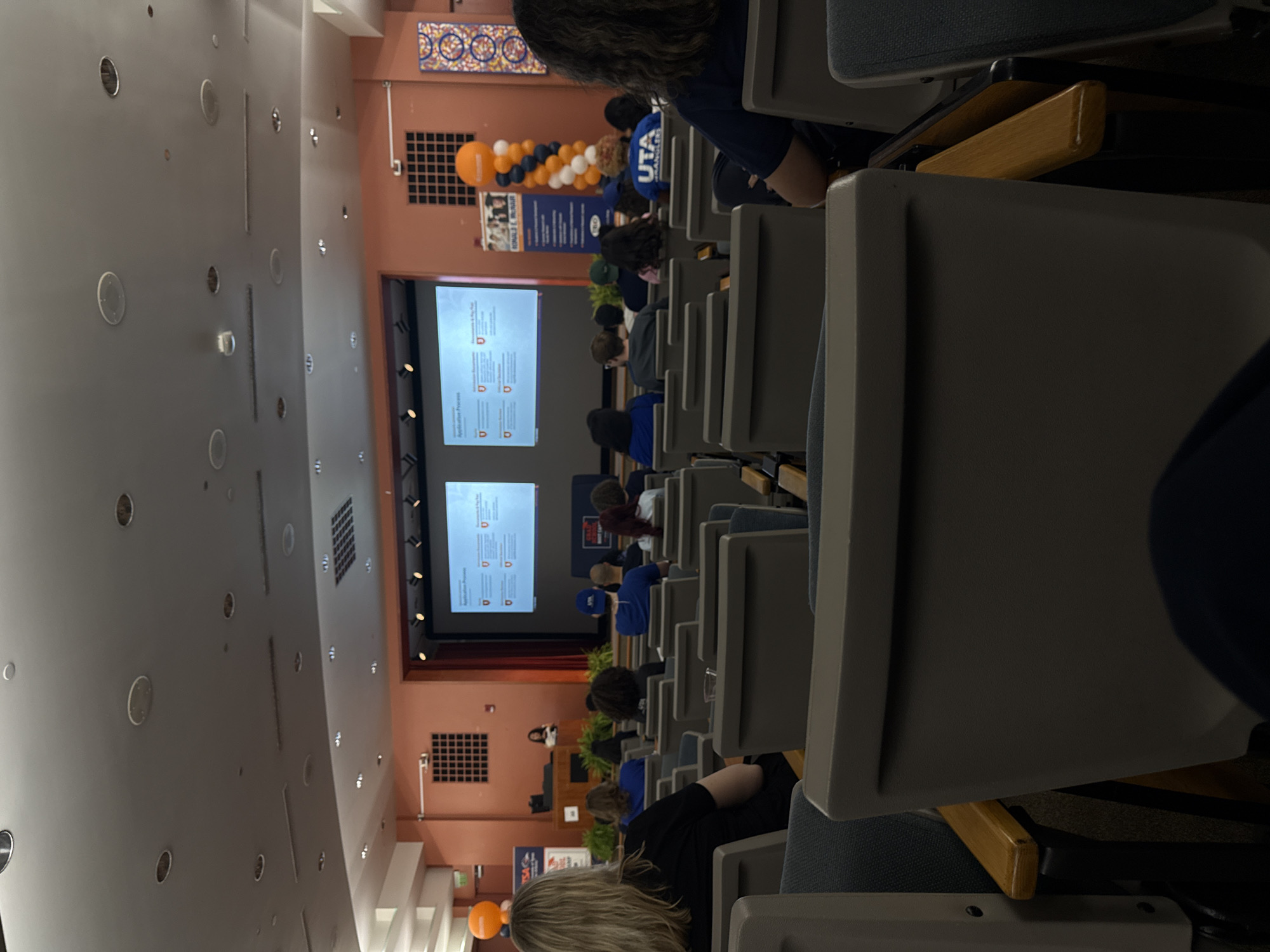 The image shows an auditorium filled with people sitting in rows of gray and blue seats with wooden arms. Two large screens at the front display information. Orange and blue balloons are visible near a promotional banner. A speaker stands at a podium on stage. The event is at the University of Texas at Arlington, although the specific location within San Antonio is not discernible from the image.
