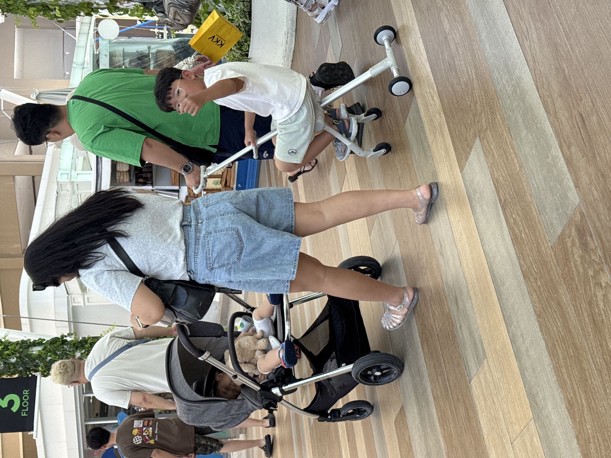 In Patong, Thailand, outside a retail establishment, a blonde-haired man in a light-colored t-shirt and a woman with long dark hair wearing a grey t-shirt and denim shorts walk. The woman pushes a dark grey stroller containing a sleeping child with a stuffed animal. To their right, a boy wearing glasses, a white t-shirt, and light-colored shorts sits on a small white riding toy. A man in a bright green t-shirt stands behind the boy. A yellow bag with the logo "KKV" is visible near the boy. The ground is tiled with a wood-grain pattern. A sign reading "3 FLOOR" is visible in the background.