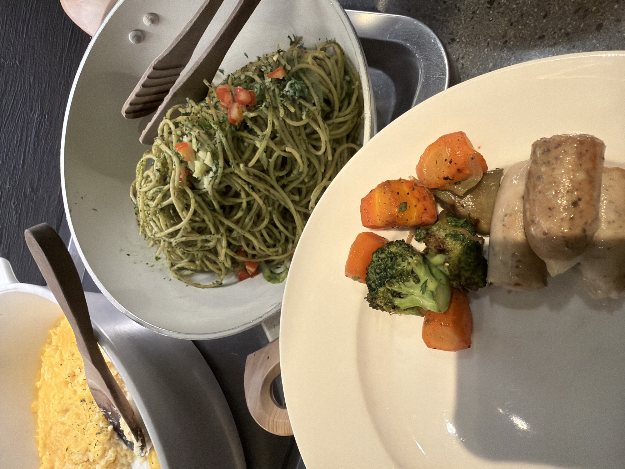 The image shows a buffet spread in a restaurant setting. In the foreground, a white plate holds an assortment of food, including roasted chicken pieces, diced carrots, and broccoli florets. To the left of the plate, a large white bowl contains what appears to be scrambled eggs, with a wooden serving spoon resting inside. Behind this, a white pot holds spaghetti with a green sauce and chopped tomatoes, with wooden tongs inserted into it. The background is dark and out of focus. The overall scene depicts a self-serve meal station.