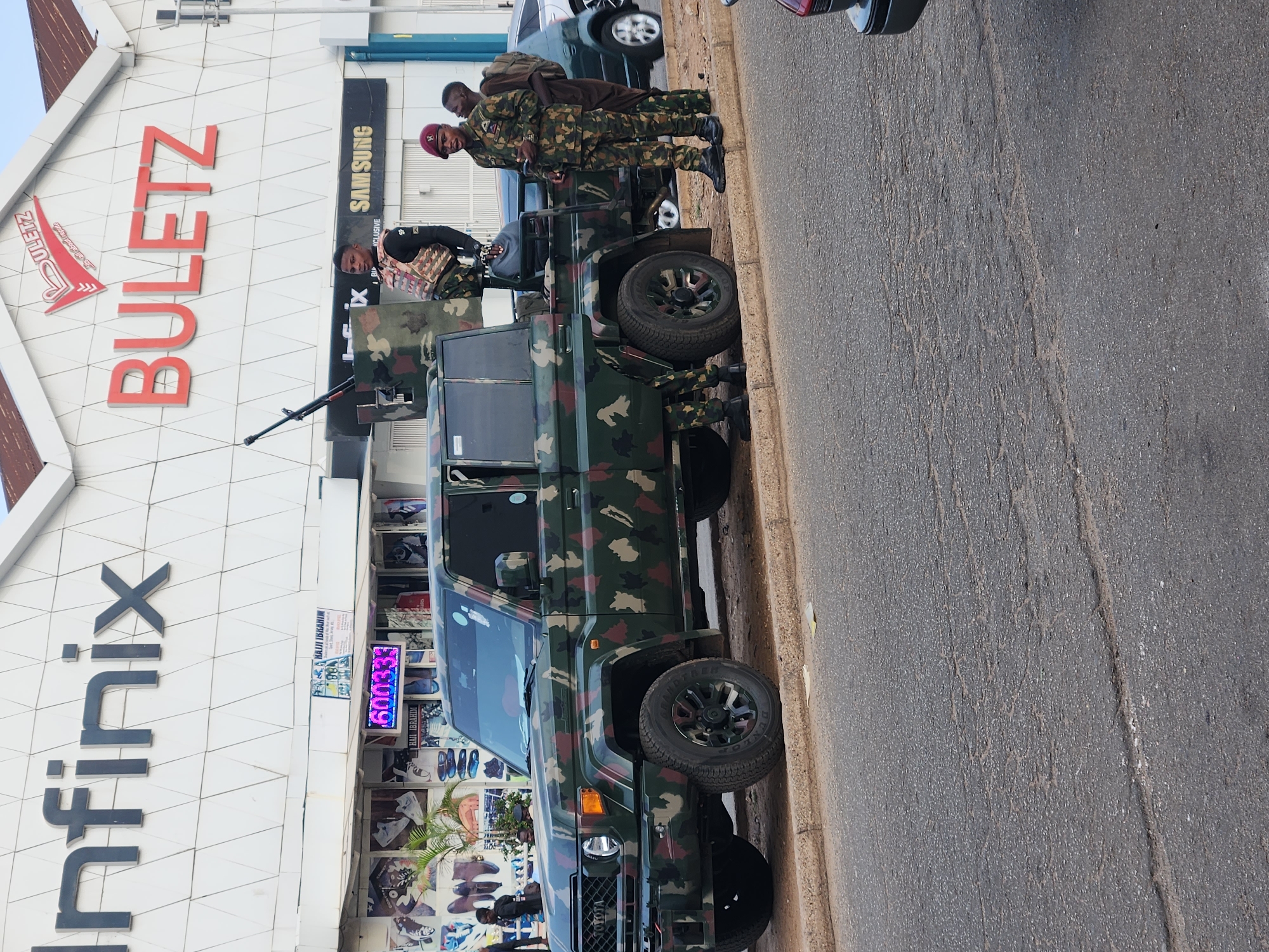 The image depicts a military vehicle with a mounted gun parked on the side of a street in Jos, Nigeria. Three men in military uniforms are standing near the vehicle. The building behind the vehicle has signage for "Infinix" and "Buletz," indicating a commercial area. A "Samsung" advertisement is also visible on the building. The street appears to be paved and shows signs of wear and tear.
