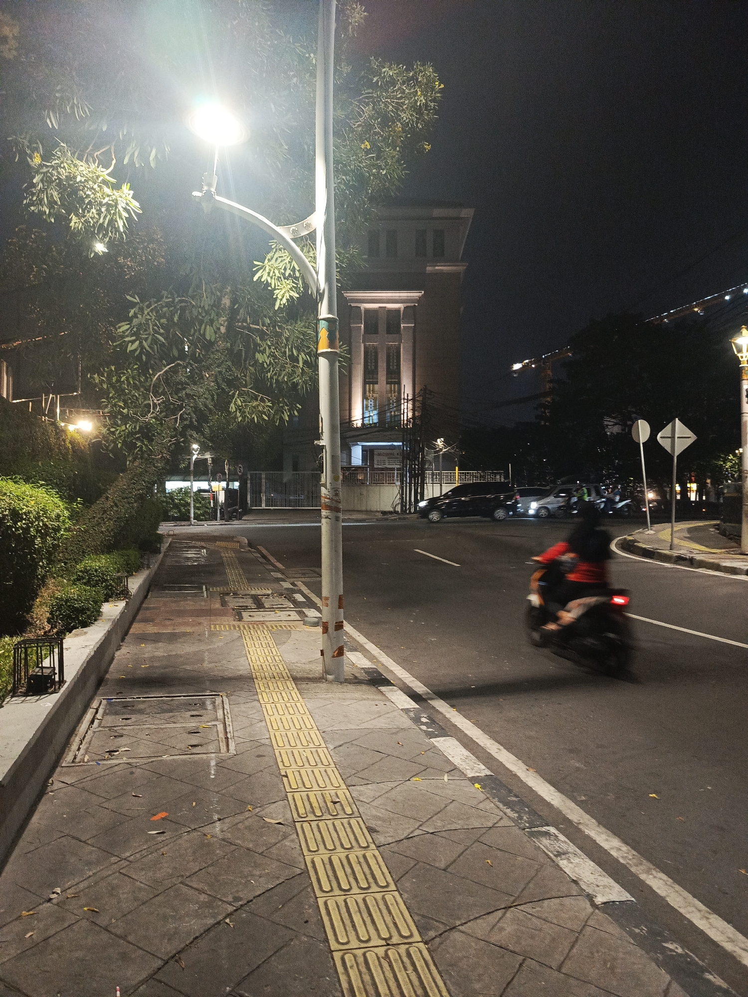 The image depicts a street scene at night in Central Jakarta, Indonesia, possibly along Jalan Sudirman. A street lamp illuminates the sidewalk, which features tactile paving for the visually impaired. Adjacent to the sidewalk is a road with traffic. A motorcycle, ridden by a person wearing a red shirt, is captured in motion on the road, its movement creating a blur. Several cars are parked or driving in the background. A multi-story building with illuminated windows stands behind the trees and sidewalk. The overall atmosphere is typical of a moderately busy urban environment at night.