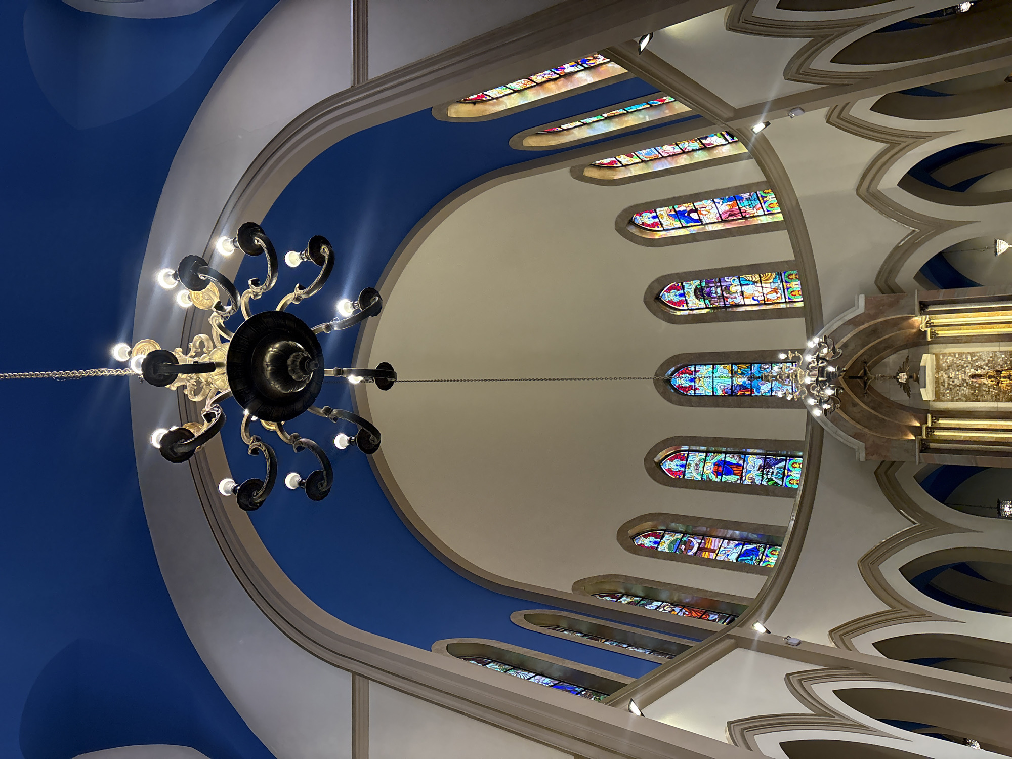 The image shows the interior of a church, likely a cathedral, with soaring arches and stained-glass windows. A large, ornate chandelier hangs from the ceiling, illuminating the space. The walls are painted white, and the ceiling is a vibrant blue. The stained-glass windows depict various religious scenes and figures in rich, colorful detail. The architecture suggests a Gothic or Neogothic style. The overall impression is one of grandeur and reverence. Without additional information, the specific church in São Paulo cannot be identified.