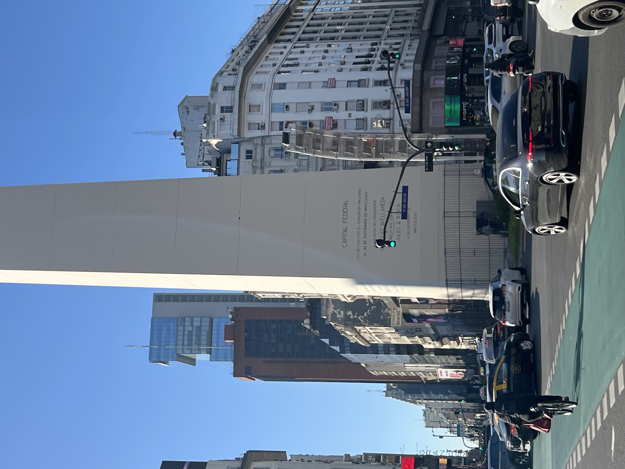 This image captures a street scene in Buenos Aires, Argentina. The Obelisco, a prominent white obelisk, dominates the left side of the frame under a clear blue sky. The monument is partially obscured by buildings. Buildings line the street on the right, with a mix of architectural styles. Traffic is moving along the street, including cars, taxis, and motorcycles. Sidewalks are visible on both sides of the street. Street signs are present, one is a blue street sign with white letters that reads "9 DE JULIO". The text on the obelisk reads, "CAPITAL FEDERAL". The scene appears to be taken during daylight hours, and the overall environment suggests a bustling urban setting.