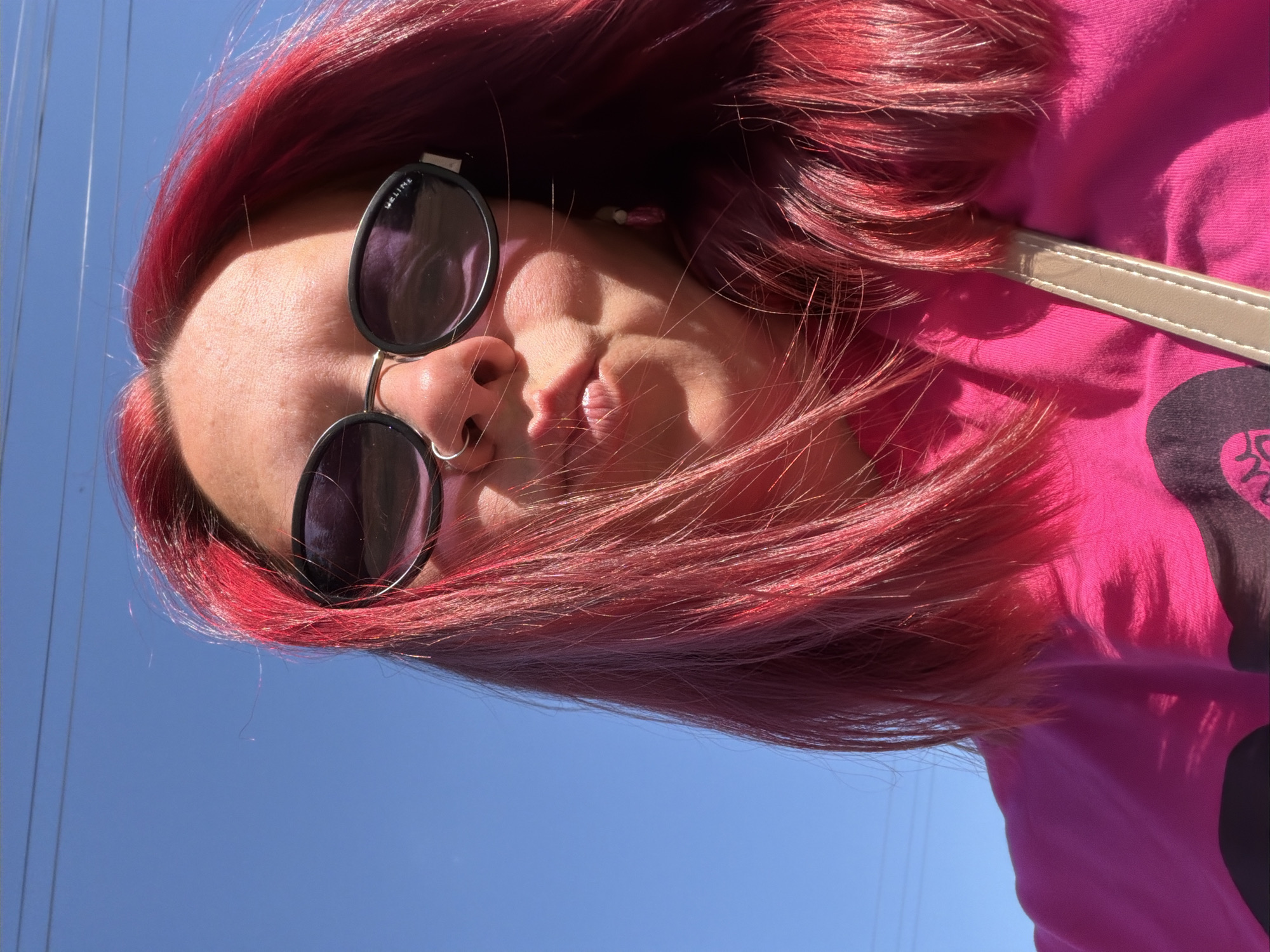 A woman with bright red hair is wearing sunglasses and a pink shirt with a graphic design. She has a nose ring. She is outdoors with a clear blue sky in the background. Power lines are visible in the sky. The location is unspecified, so no information about Chaikovsky, Russia, can be inferred from the image.