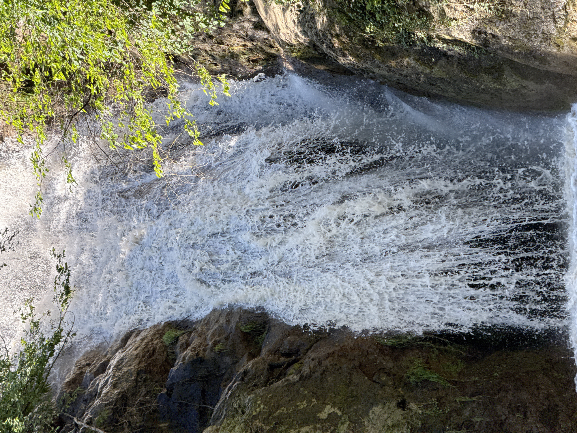 A view from above looking down at the Hotnitsa Waterfall in Hotnitsa, Bulgaria. Water cascades over a rocky cliff, creating a white, foamy texture in the center of the image. Vegetation, including trees and moss, grows on and around the rocks. The waterfall flows down a rocky cliff face, and the presence of trees on the left side and in the foreground adds a natural element to the scene. The water's motion creates a dynamic and textured effect.