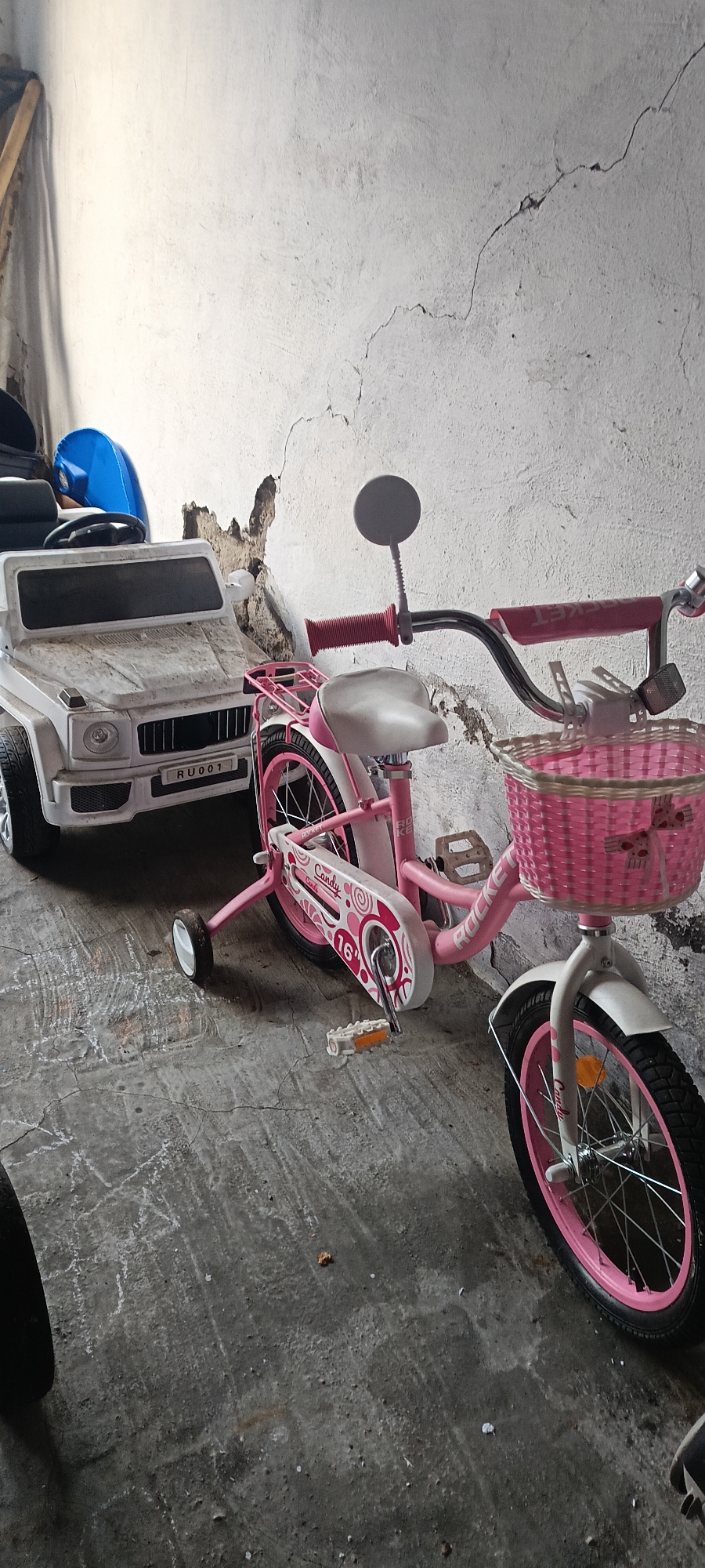 In Хохлы, Russia, within an unlit, cracked-wall garage, a white toy car with a license plate reading "RU001" is parked next to a pink children's bicycle. The bicycle features training wheels, a pink wicker basket on the handlebars, and the word "Candy" written on its frame. The floor of the garage is a rough concrete surface with visible chalk marks. The overall scene suggests a storage area for children's toys.