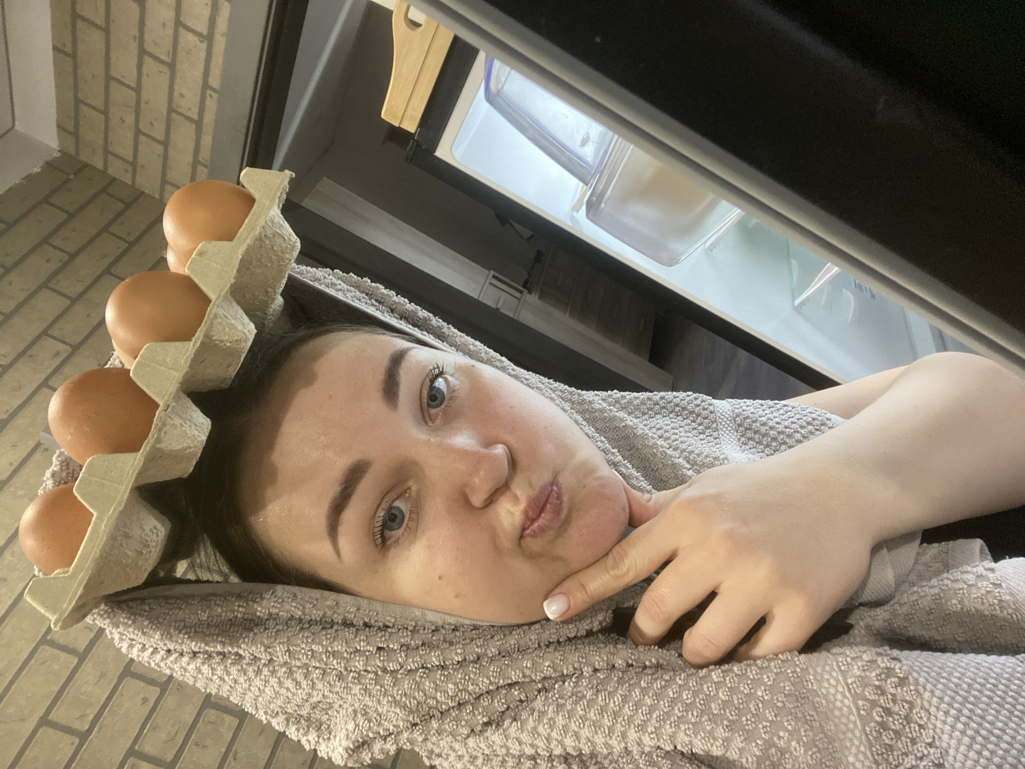 A woman lies on her side on a grey towel with an egg carton containing four brown eggs balanced on her head. She looks directly at the camera with pursed lips and touches her chin with her right index finger. The open door of a refrigerator is visible in the background, revealing shelves and containers inside. The floor is covered in brick tiles. The image was not taken in Sochi, Russia.