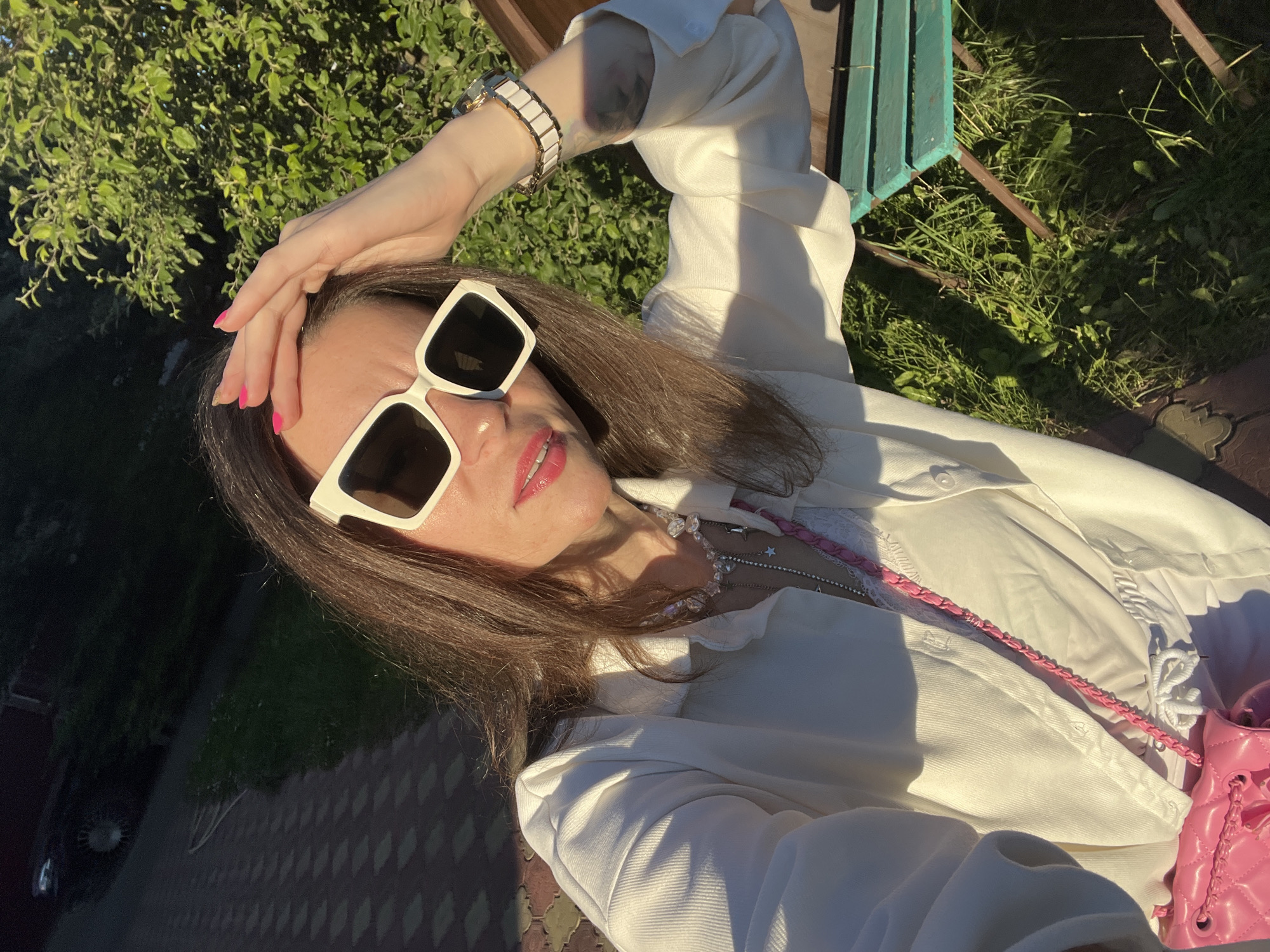 The media file shows a medium shot of a woman posing for a selfie outdoors. She is wearing a white shirt, white sunglasses with thick frames, and several necklaces. Her hand is resting on her head, and she is smiling. A pink quilted bag with a chain strap is visible. The background includes greenery, a brick pathway, and a portion of a turquoise and brown bench. The lighting suggests it is daytime, possibly late afternoon, with the sun casting distinct shadows, indicating fair weather. The woman's facial expression and pose convey a relaxed and cheerful emotion. The location context is Krasnoe-na-Volge, Russia. No visible text is present in the image.