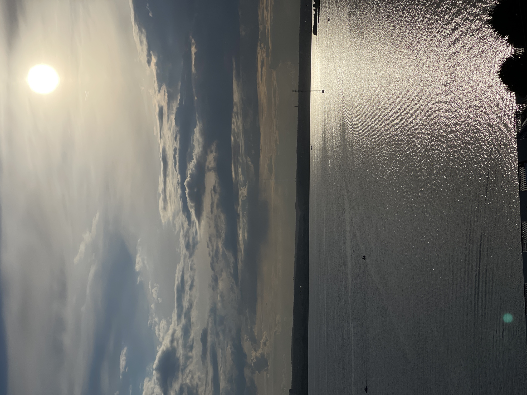 The image depicts a body of water reflecting the bright sun, creating a shimmering effect with concentric ripples. The sun is positioned in the upper left quadrant of the image, casting a warm glow. Dark clouds are visible in the upper portion of the sky, contrasting with the sunlit haze. A distant shoreline is visible across the water, with silhouetted structures that might include buildings and a tall mast. The overall scene suggests an outdoor environment, possibly a lake or ocean, during daylight hours. The water is calm with gentle waves. There are no discernible people or specific objects in the water except for a few small, dark shapes that could be boats or debris. The mood is serene and tranquil, with the natural light dominating the composition. The image is oriented vertically.