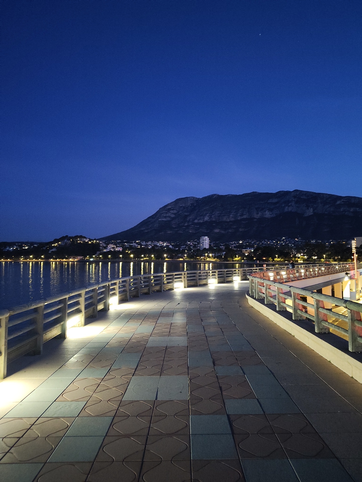 The image displays a nighttime view of a paved promenade in Dénia, Spain. The promenade, constructed with alternating light brown and blue square tiles featuring an embossed curvilinear pattern, extends from the foreground and curves subtly to the right. Integrated linear ground lights illuminate the pathway and the base of a light-colored, slatted railing bordering the left side of the promenade. A similar railing, incorporating reddish elements, is visible along the right edge. To the left, a calm body of water reflects numerous bright yellow and white lights from the distant city across the bay. The background reveals the illuminated urban landscape of Dénia, with buildings and streetlights, set against the dark, prominent silhouette of the Montgó mountain massif. The sky above is a deep indigo, with a faint crescent moon and a few stars visible in the upper right quadrant. No individuals are present in the scene.