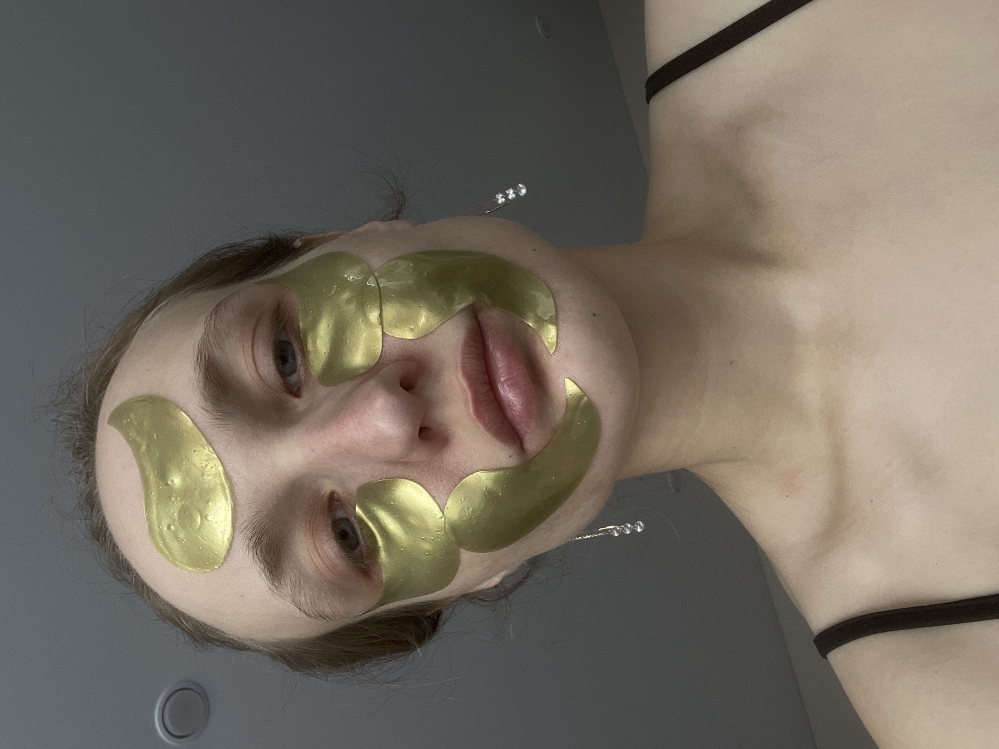 A fair-skinned person lies on their back, looking up towards the camera. They have light eyes, light hair, and are wearing gold-colored facial patches arranged around their eyes and mouth. One patch is on their forehead, two under each eye, and two above the upper lip. They have jewelry in their ears. The background is a plain gray. They are wearing a black top with thin straps. The image appears to have been taken indoors. There is no information in the image to suggest this photo was taken in Подшивалово, Russia.