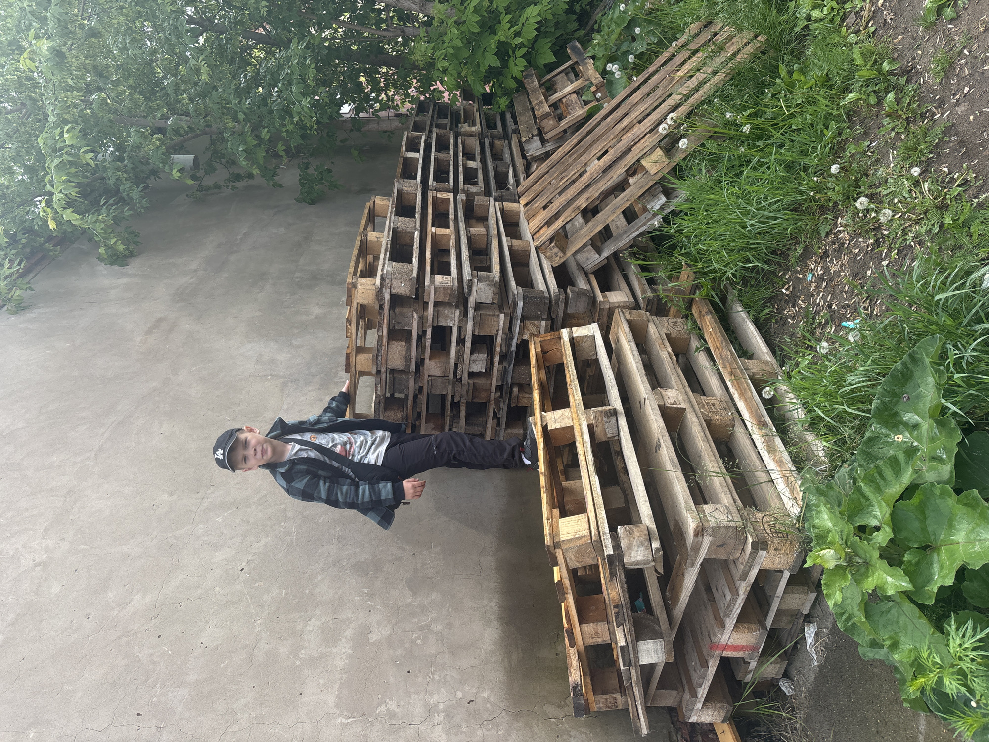 The image shows a young boy standing outdoors next to a large stack of wooden pallets. The boy, who appears to be pre-teen, is wearing a black baseball cap with a white "LA" logo, a dark plaid long-sleeve shirt over a light-colored t-shirt, dark trousers, and dark sneakers with white and red accents. He is leaning slightly, with his left arm resting on the stack of pallets, and looks directly at the camera with a neutral to slightly smiling expression.
The main subjects in the scene are the boy and the substantial collection of wooden shipping pallets. These pallets are stacked in tall piles, primarily to the boy's right and behind him. Many of the pallets show signs of wear and weathering. On one of the pallets, the letters "EPA" are clearly visible.
The setting appears to be an outdoor area, possibly a backyard or a storage space in Chaikovsky, Russia. The ground beneath the boy is a cracked concrete or asphalt surface. To the right of the pallets, the ground transitions into dirt and wild vegetation, including green grass, white dandelions, and larger broad-leafed plants. Overhead and in the background, lush green tree foliage is visible.
The activity taking place is the boy posing for a photograph. The time of day appears to be daytime, likely overcast, as the light is diffused, and there are no strong shadows.