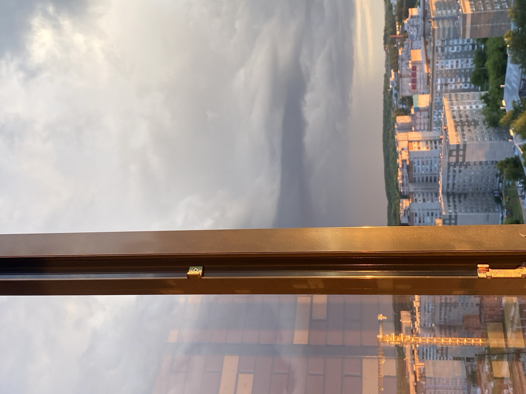 The image shows a view from a high vantage point, likely a window, overlooking a cityscape. The foreground features a metal window frame with visible hardware. The midground is split between a cloudy sky and a cityscape with multi-story residential buildings, interspersed with trees. The buildings are a mix of white, gray, and red-brown. The reflection in the glass of the window shows a distorted view of some buildings and a patterned surface, likely the floor or wall the window is adjacent to. The photograph is rotated horizontally.
It is difficult to determine the location with certainty.