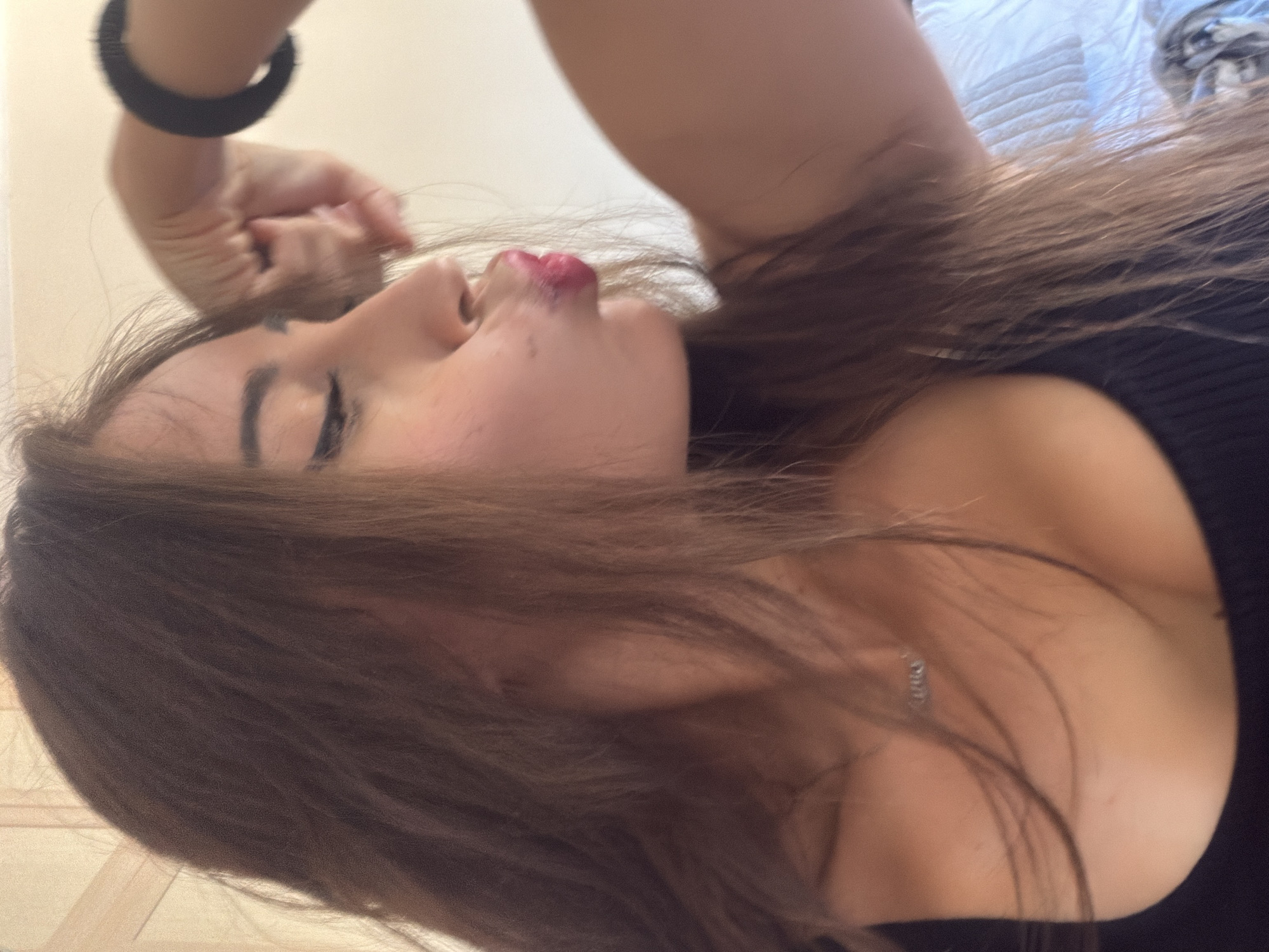 In a close-up, profile view, a young woman with long, brown hair is captured making a kissing face, her eyes closed. She is wearing a black top, and a delicate silver necklace with a small charm is visible at her collarbone. Her right arm is raised, and her fist is clenched, adorned with a black wristband. The background consists of a plain, light-colored wall and a white, textured surface that may be a bedspread. The lighting suggests an indoor setting. The scene is set in Tersefanou, Cyprus.